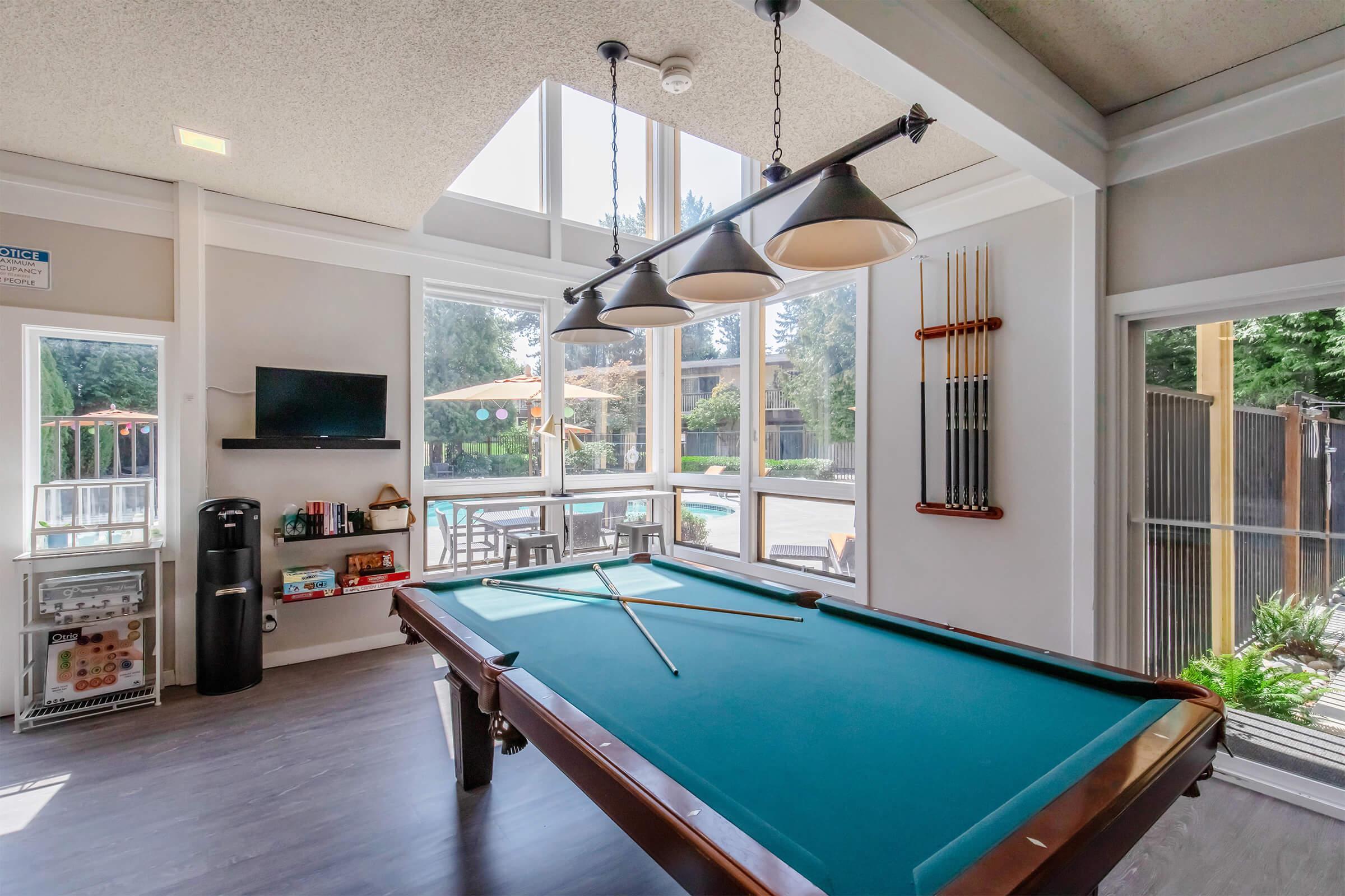 A bright and spacious game room featuring a green pool table with a cue stick. Large windows allow natural light to flood in, showcasing an outdoor pool area. The room includes a flat-screen TV, bookshelves, and seating arrangements by the windows for relaxation.