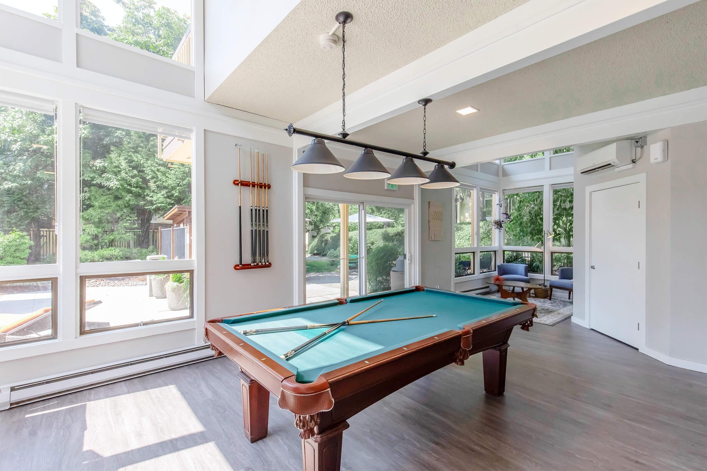A bright and airy game room featuring a billiards table with green felt, surrounded by large windows that offer views of the outdoor space and lush greenery. The room has modern lighting fixtures and comfortable seating, creating a welcoming atmosphere for leisure and entertainment.