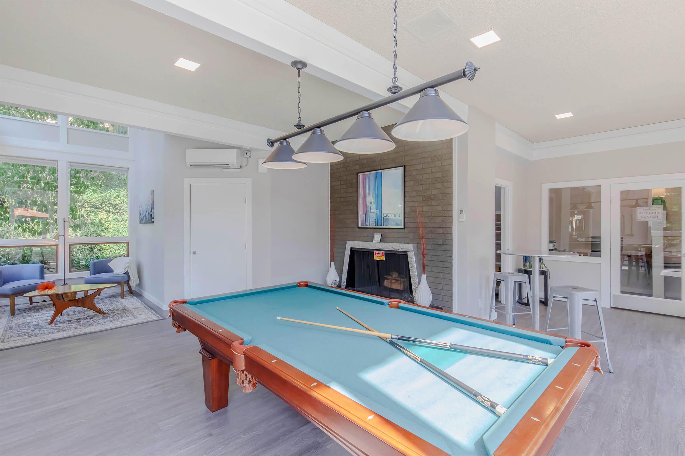 A brightly lit game room featuring a billiards table with cue sticks, surrounded by modern decor. There are large windows providing natural light, a cozy seating area with a rug, and a fireplace. The room has a contemporary design with a light color palette and stylish light fixtures.