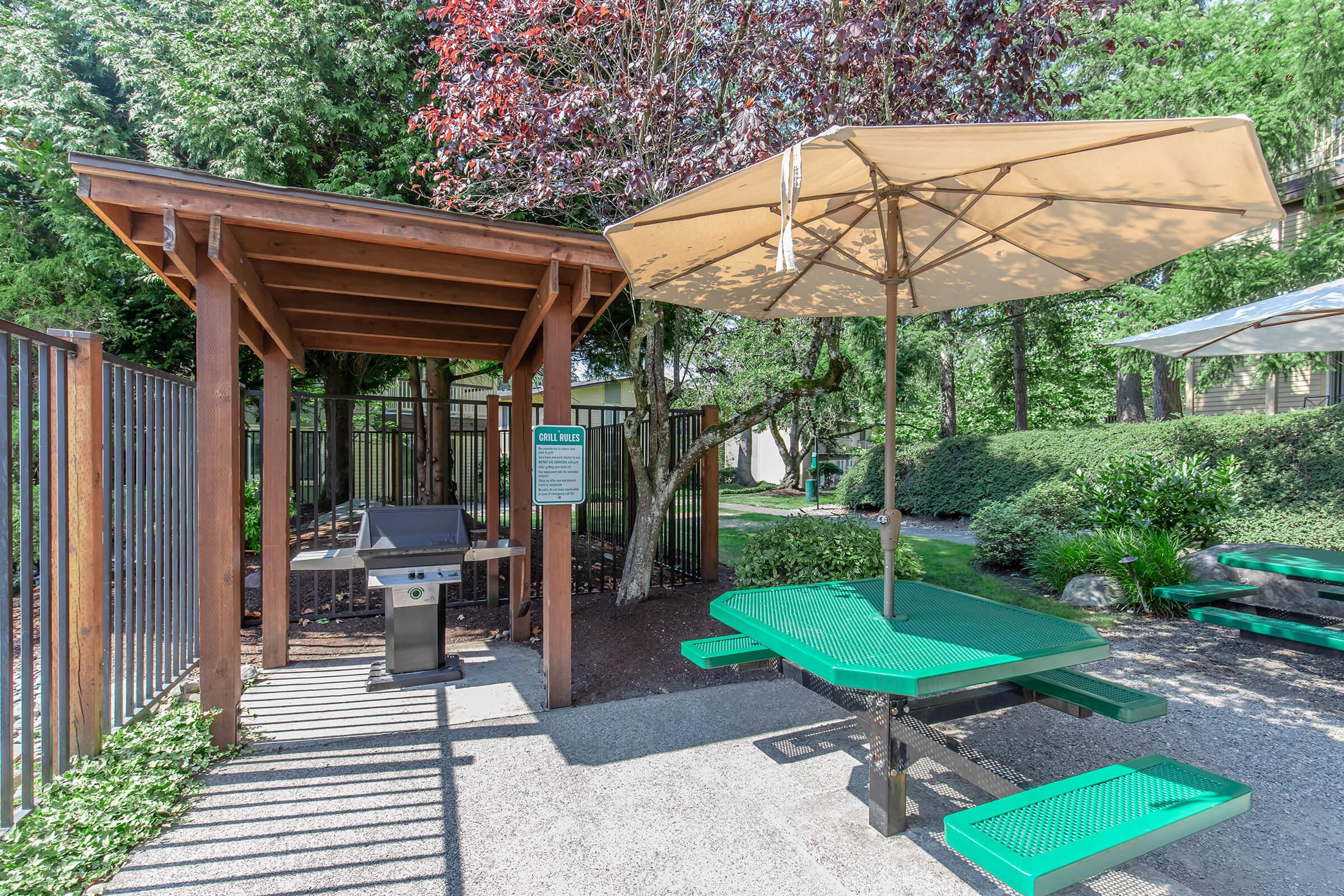 An outdoor grilling area featuring a green picnic table and a grill beneath a wooden shelter. The space is shaded by a large umbrella and surrounded by greenery, with a fence in the background. A sign is visible near the grill, providing information about the area.