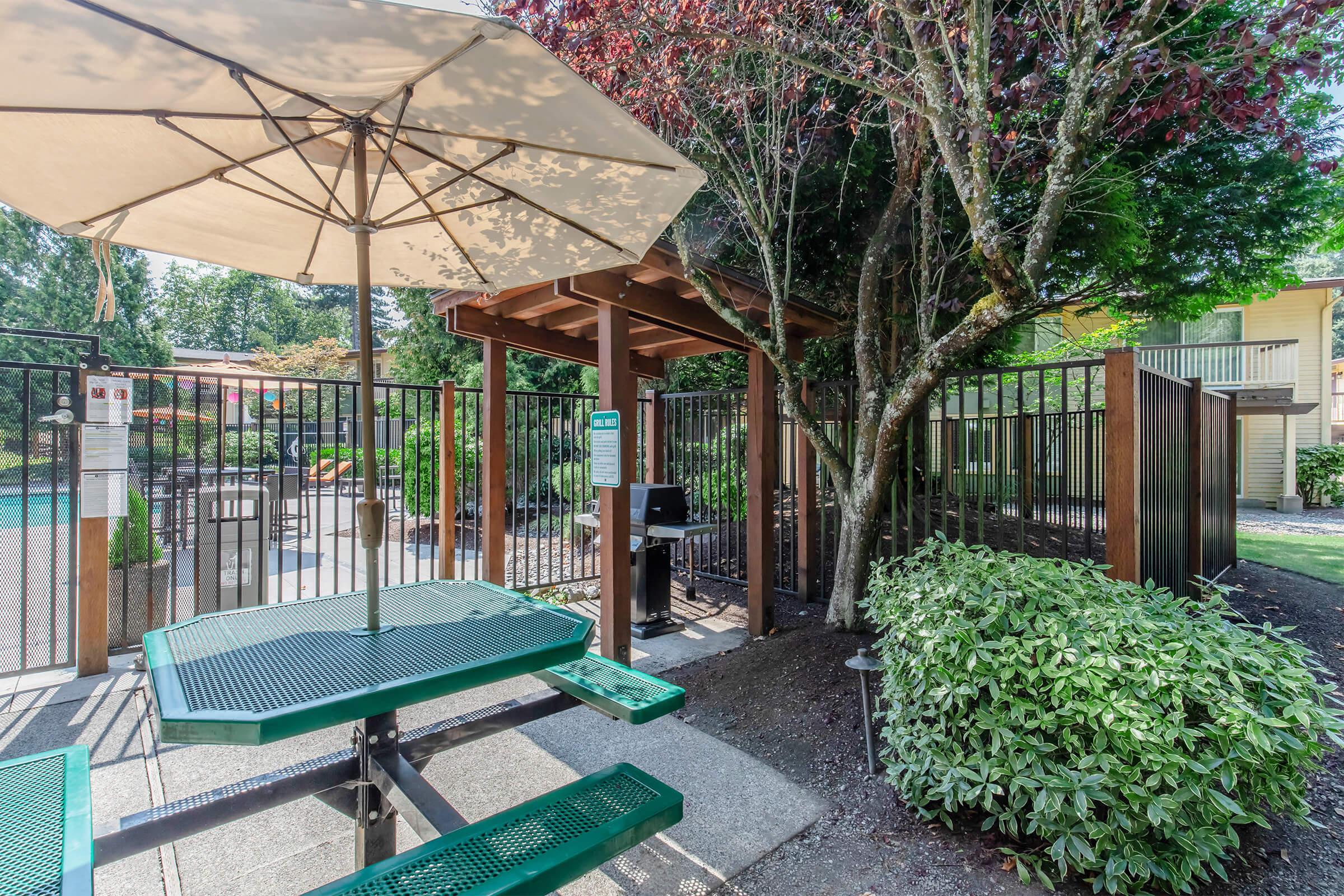 A shaded picnic area featuring a green picnic table under a large umbrella, surrounded by lush greenery. In the background, a swimming pool area is visible, enclosed by a black fence. The scene is well-lit, suggesting a pleasant outdoor setting.