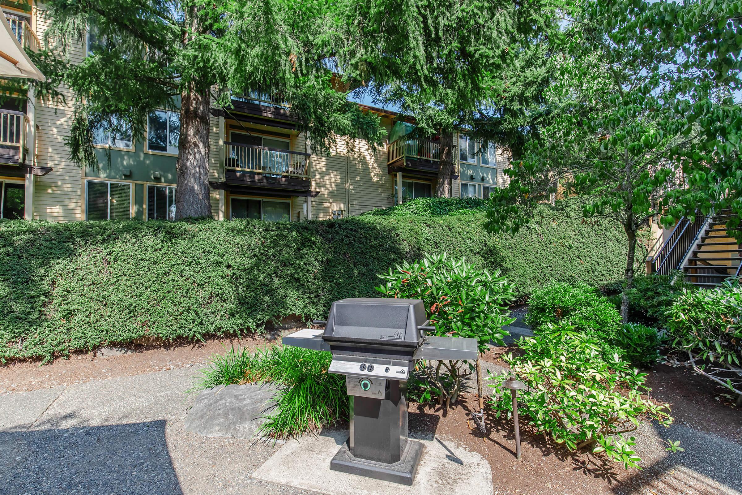 A charcoal grill stands in a landscaped outdoor area surrounded by lush greenery and shrubs, with apartment buildings visible in the background. The scene is bright and inviting, perfect for outdoor cooking and gatherings.