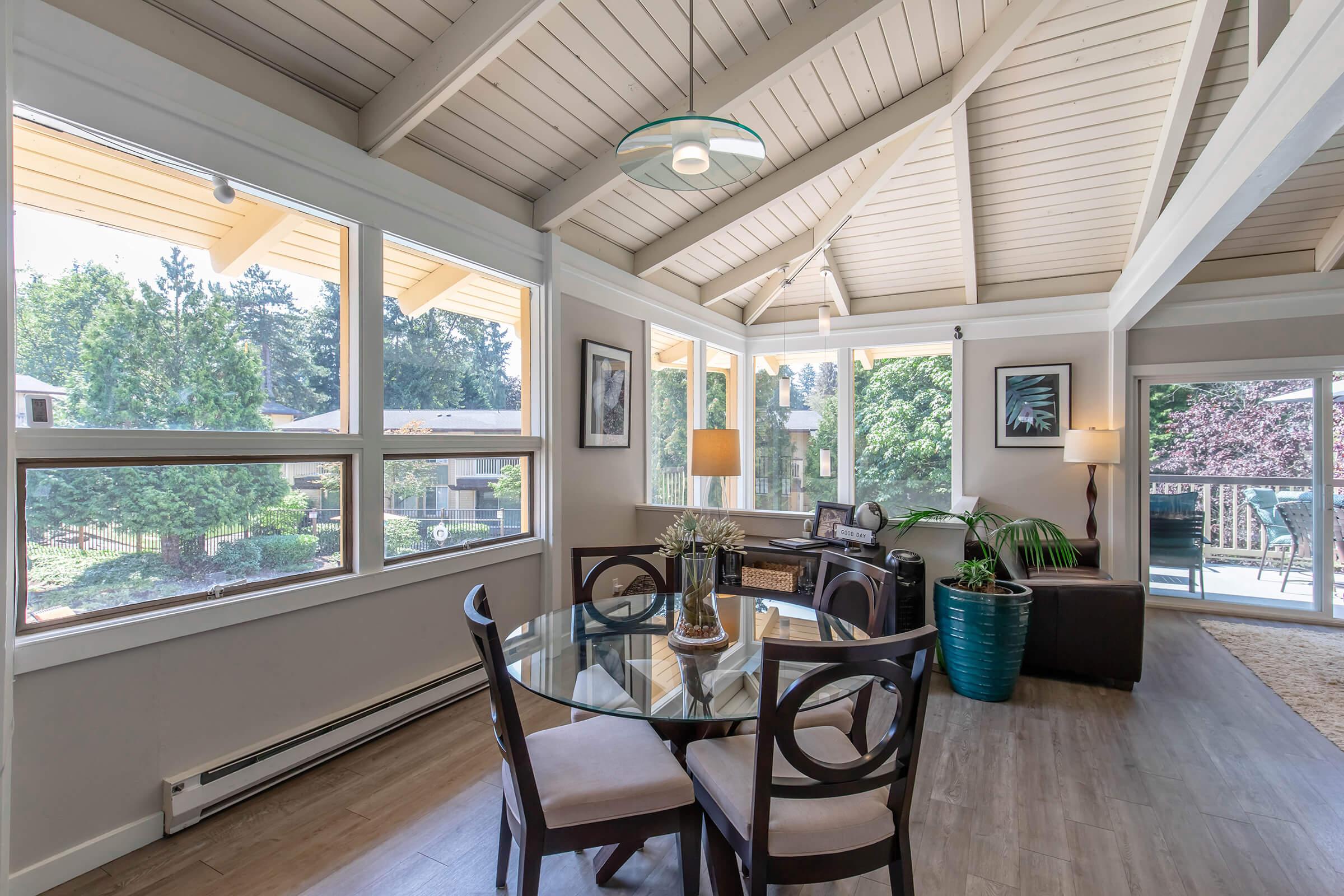 A bright and airy dining area with a round glass table and four chairs. Large windows let in plenty of natural light, showcasing the lush greenery outside. The room features vaulted wooden ceilings and a modern light fixture, along with a potted plant and decorative elements.