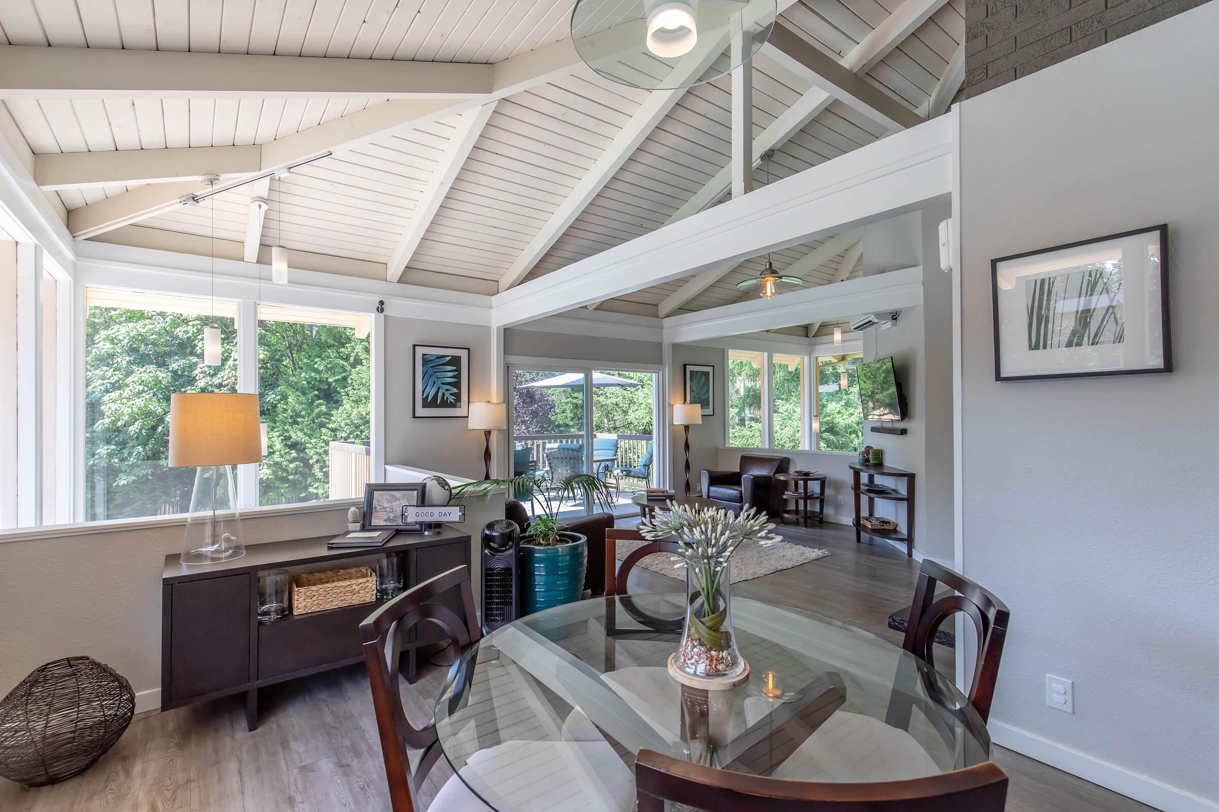 Bright and modern living space featuring a dining table with four chairs, large windows allowing natural light, a stylish lamp, and a decorated corner with a plant. The ceiling has a unique design, and there are cozy areas for relaxation, creating an inviting atmosphere.