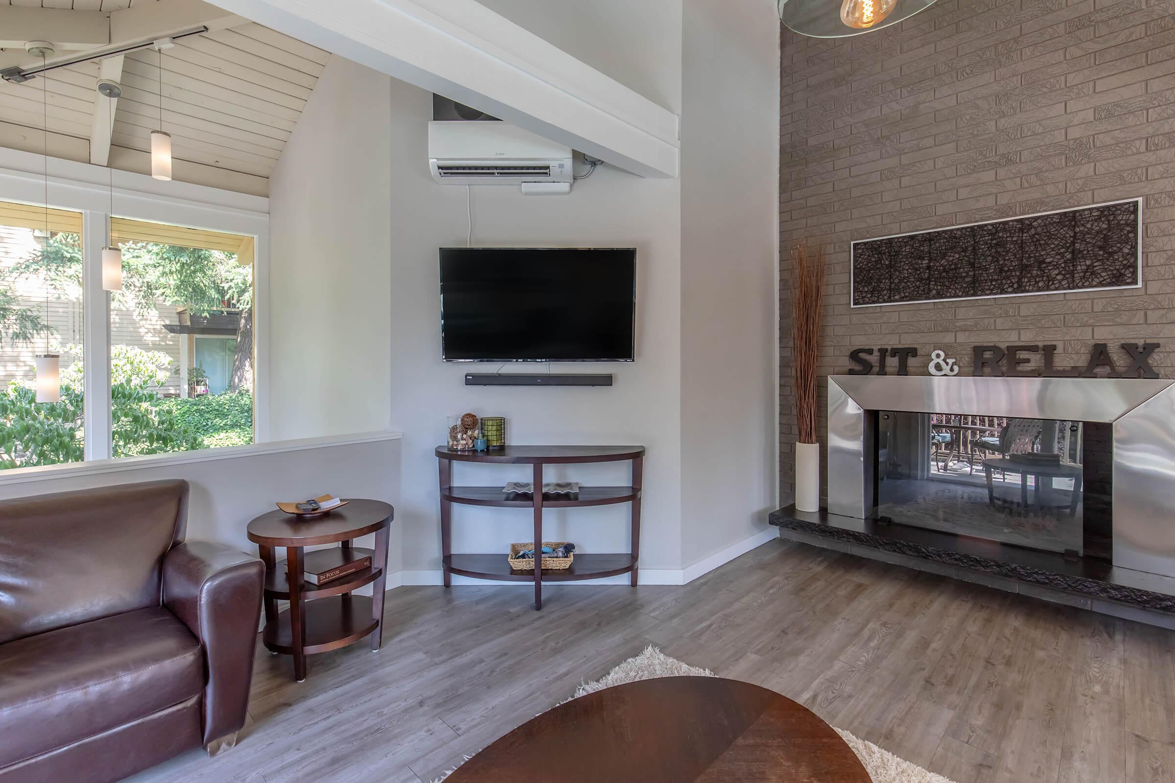 A cozy interior living space featuring a brown leather sofa, a round side table with decorative items, and a mounted television. There's a sleek fireplace with a modern design, and a wall decoration that reads "SIT & RELAX." Large windows allow natural light, enhancing the inviting atmosphere.