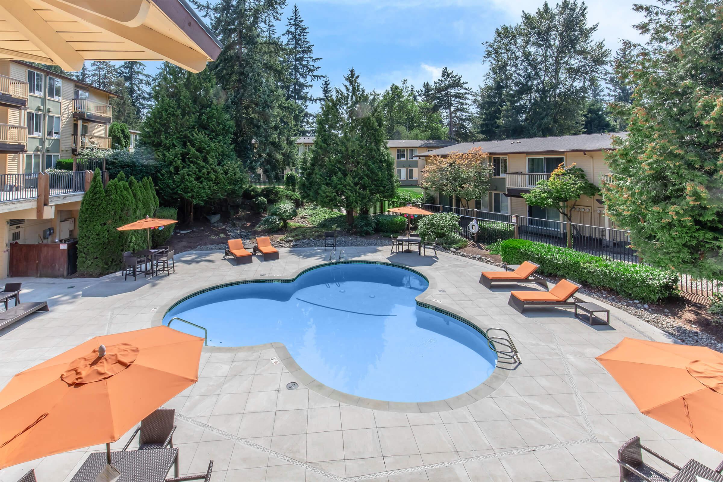 A heart-shaped pool surrounded by lounge chairs and orange umbrellas, set in a landscaped area with trees and greenery. Nearby buildings provide accommodation, creating a relaxing outdoor space. Bright blue sky with a few scattered clouds enhances the sunny atmosphere.