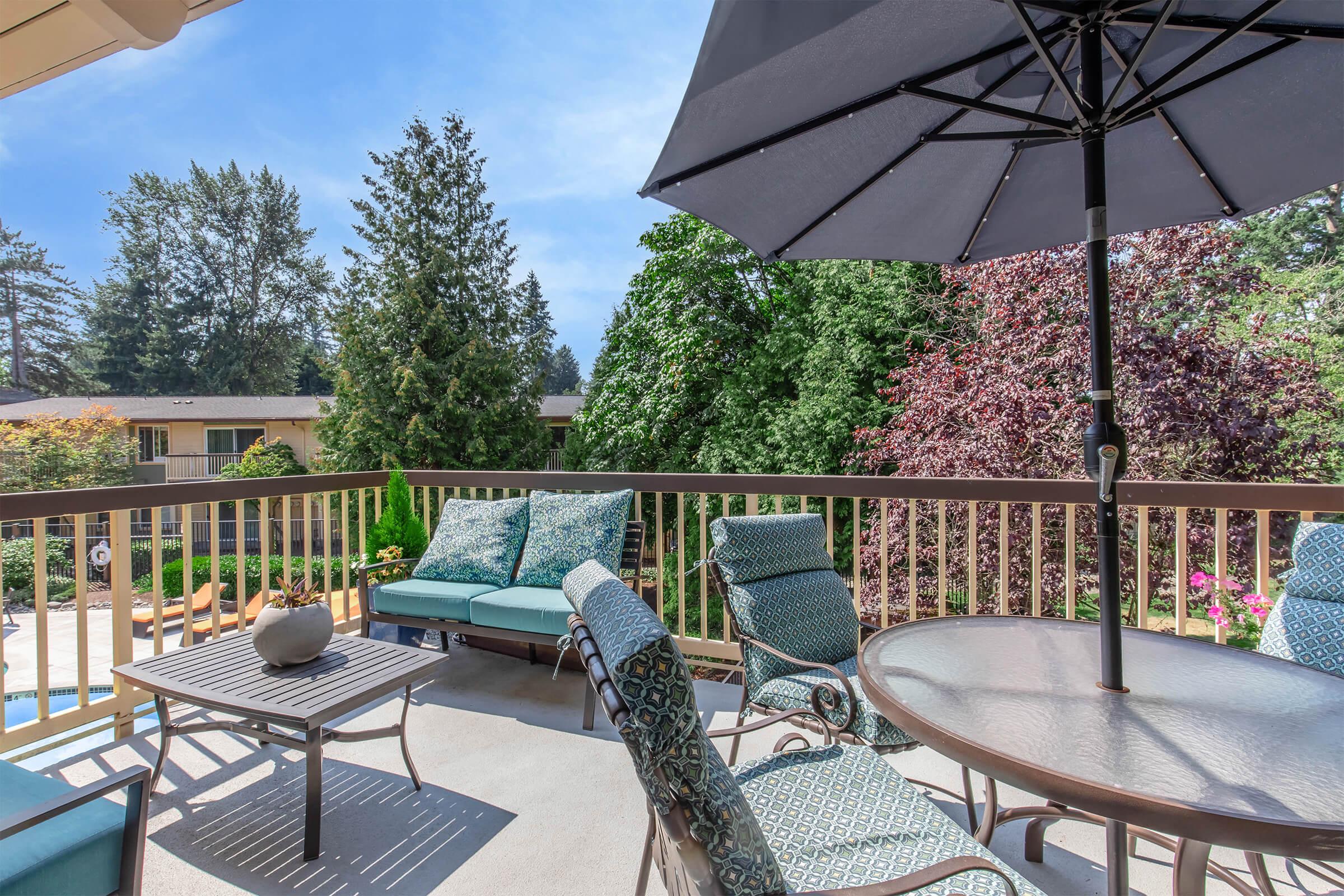 A cozy patio scene featuring a round table with chairs, a large umbrella, and turquoise cushions. Lush greenery can be seen in the background, creating a peaceful outdoor space. A pool is visible nearby, enhancing the inviting atmosphere of the deck.
