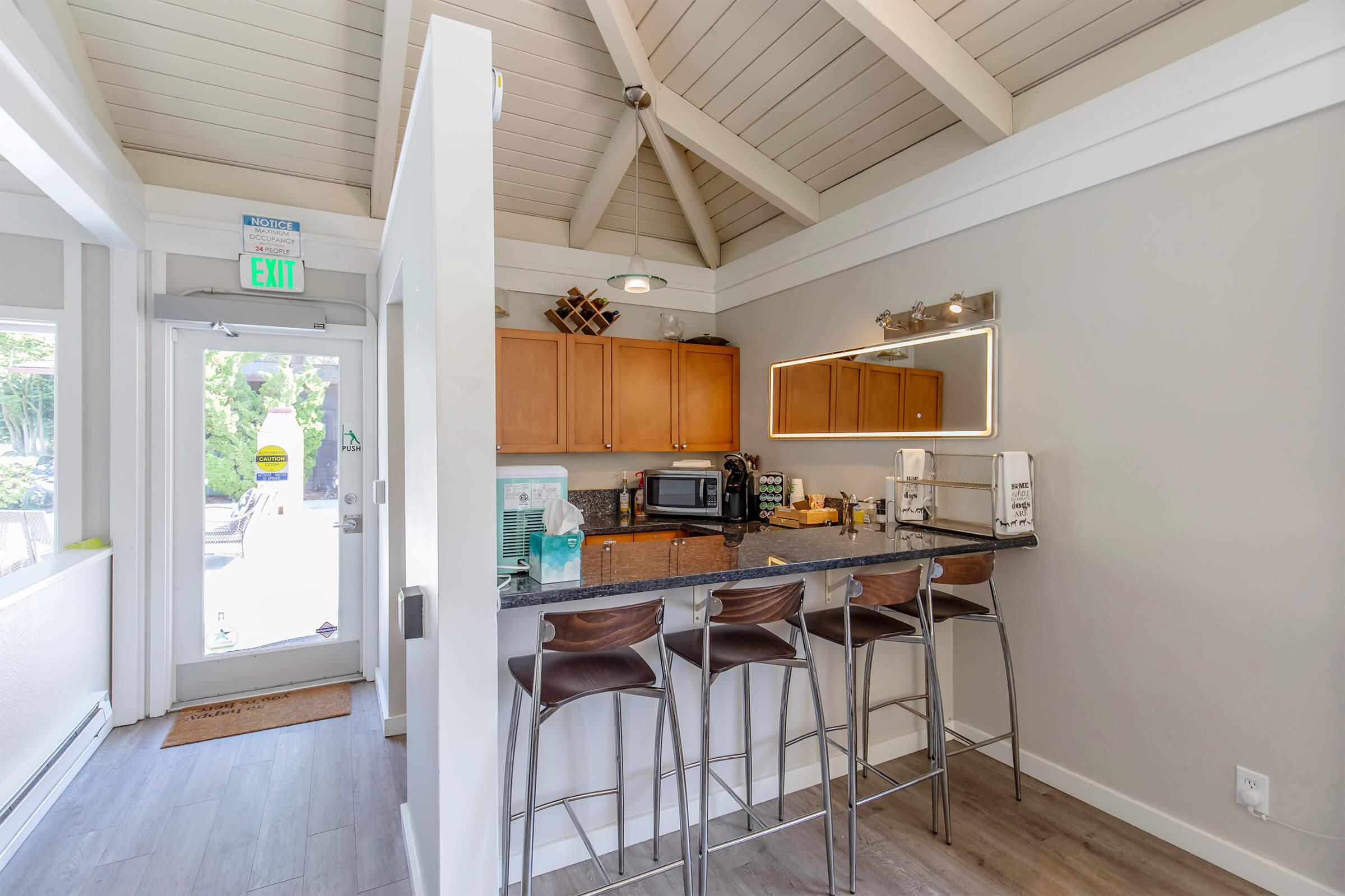 Modern kitchen area with dark wood cabinets, a black countertop, and stainless steel appliances. A bar with four high-backed chairs is situated near the counter. The space features a vaulted ceiling with wooden beams, and natural light floods in from a nearby door leading outside.