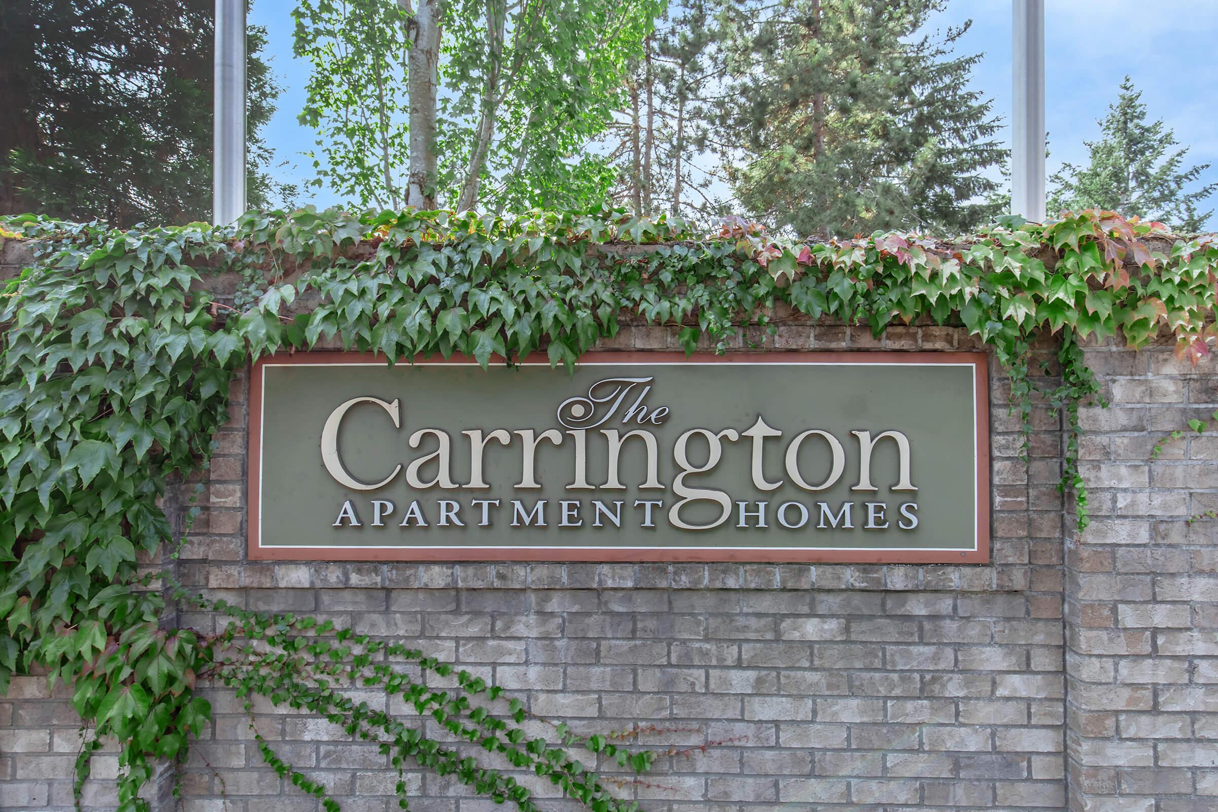 Sign for "The Carrington Apartment Homes" displayed on a brick wall, surrounded by green ivy and trees in the background. The atmosphere is inviting and well-maintained, reflecting a welcoming residential community.