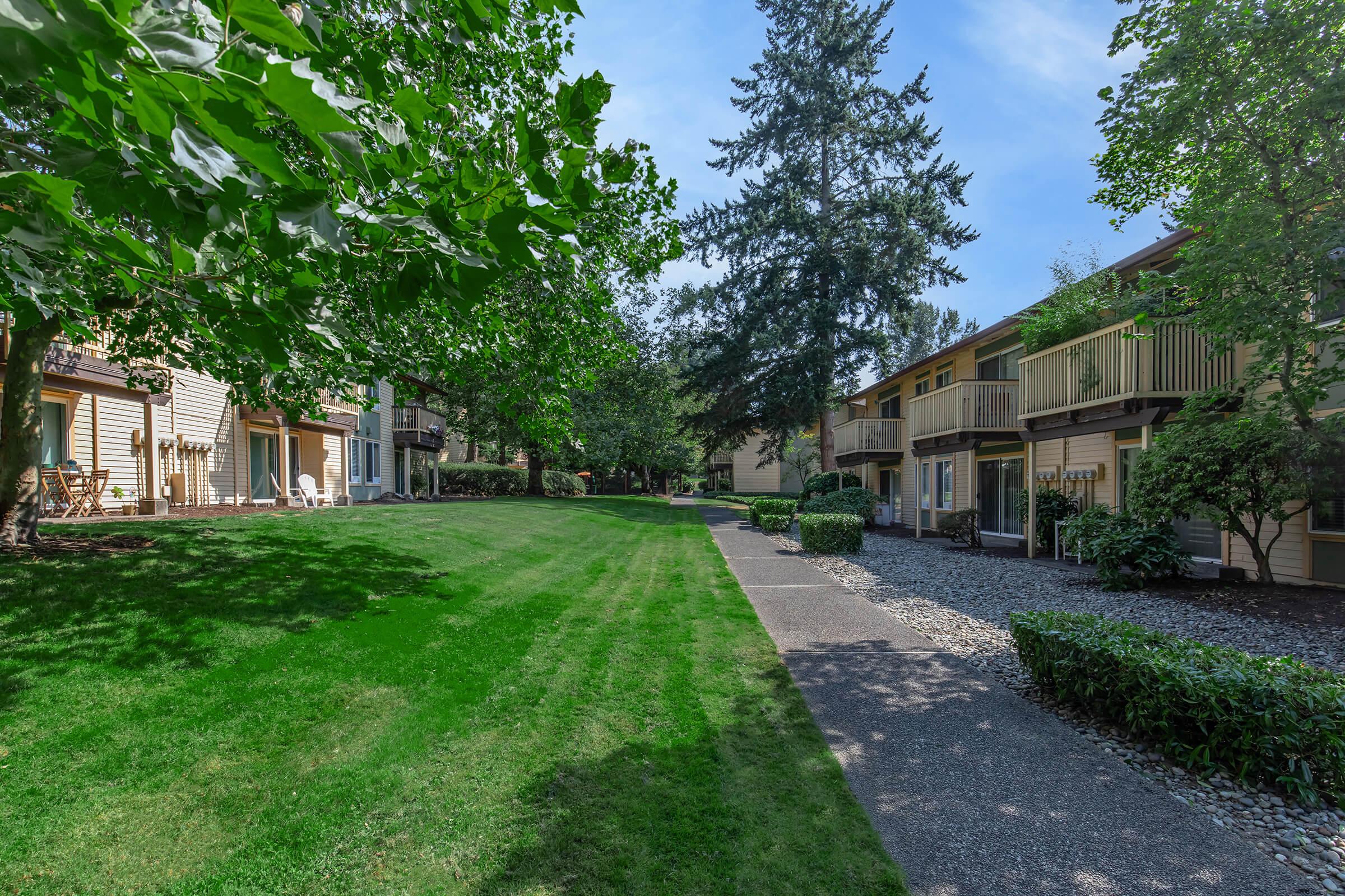 Lush green lawn surrounded by residential buildings, featuring trees and bushes. Pathway leads through the manicured landscape, showcasing a peaceful and inviting atmosphere in a well-maintained apartment complex. Bright blue sky adds to the serene setting.