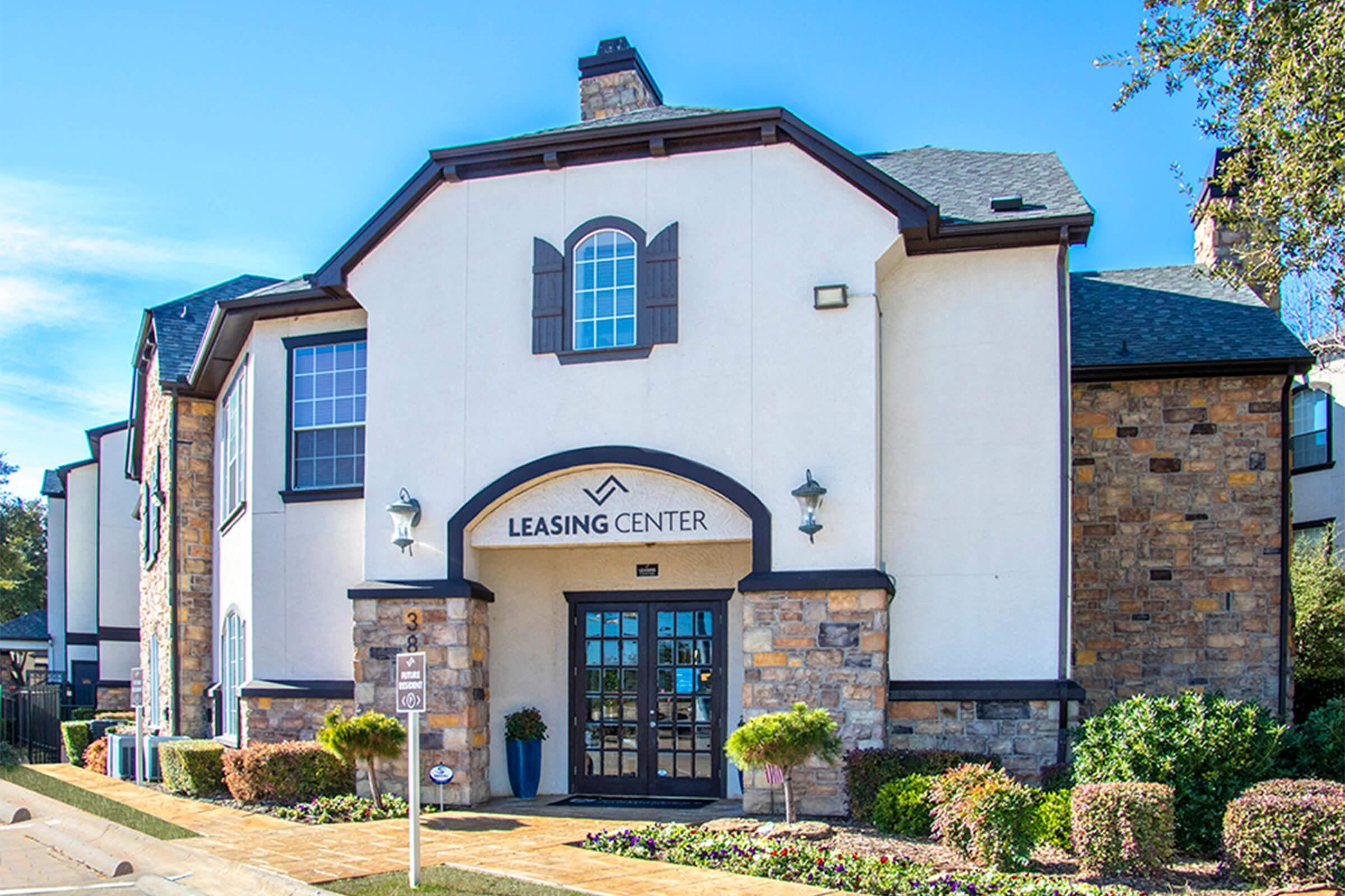 A well-maintained leasing center building featuring a combination of white stucco and stone exterior. It has multiple windows, a prominent arched entrance labeled "LEASING CENTER," and landscaped greenery in front. The architecture reflects a modern style set against a clear blue sky.