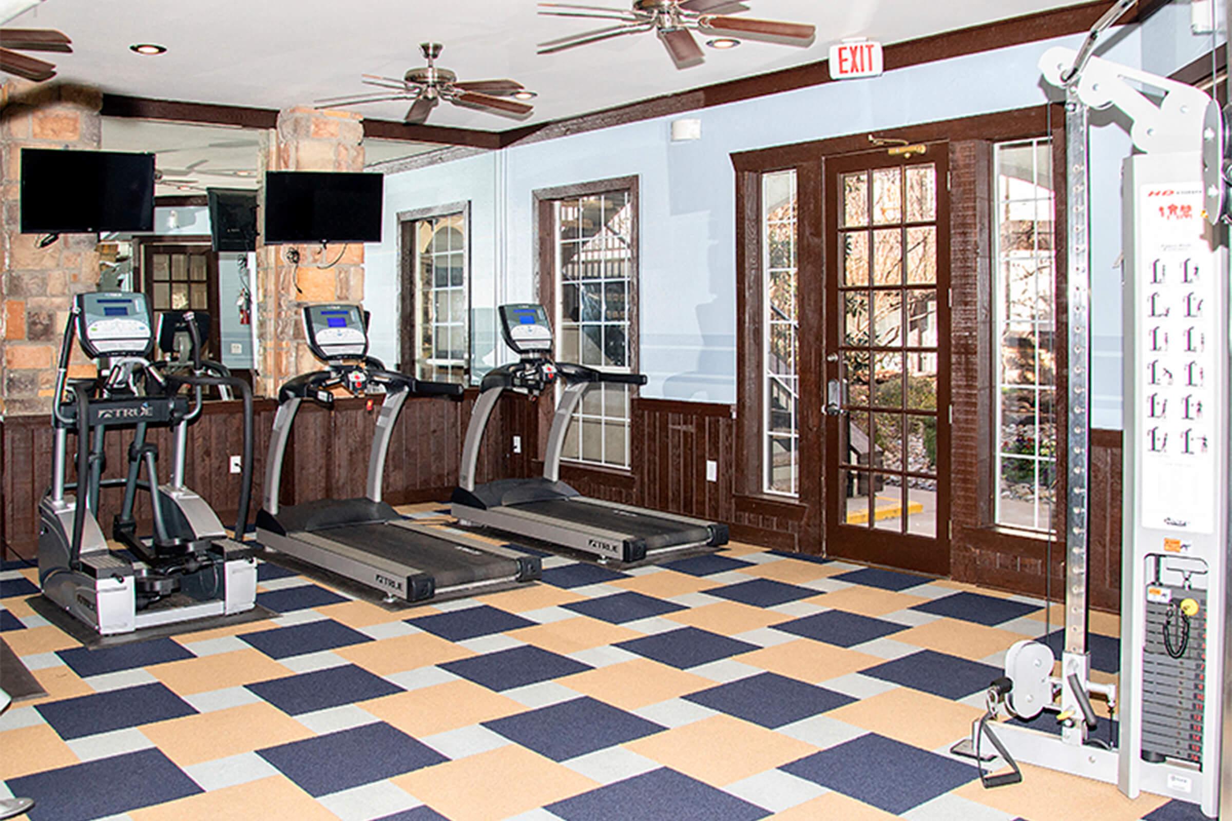 A modern gym interior featuring three treadmills, a multi-station weight machine, and two wall-mounted televisions. The floor is covered with blue and orange tiles, and there are large windows and a glass door leading outside, providing natural light to the space.
