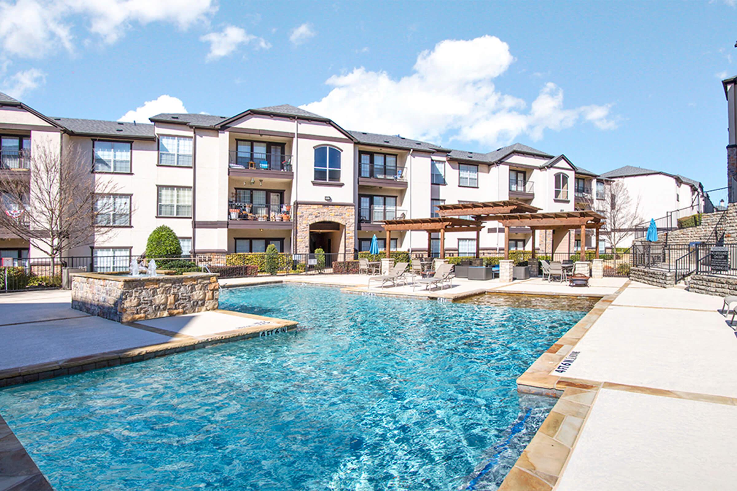 A sparkling outdoor swimming pool surrounded by a stone patio and lounge chairs. In the background, a modern apartment complex with balconies and a pergola provides a welcoming setting under a clear blue sky.