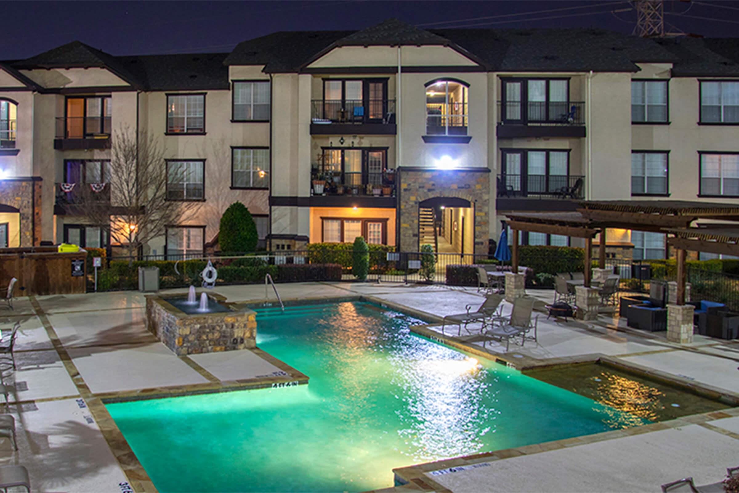 An apartment complex at night featuring a well-lit swimming pool surrounded by lounge chairs. The pool area includes a small water feature, and the building has multiple balconies with lights on. There are trees and a shaded seating area nearby.