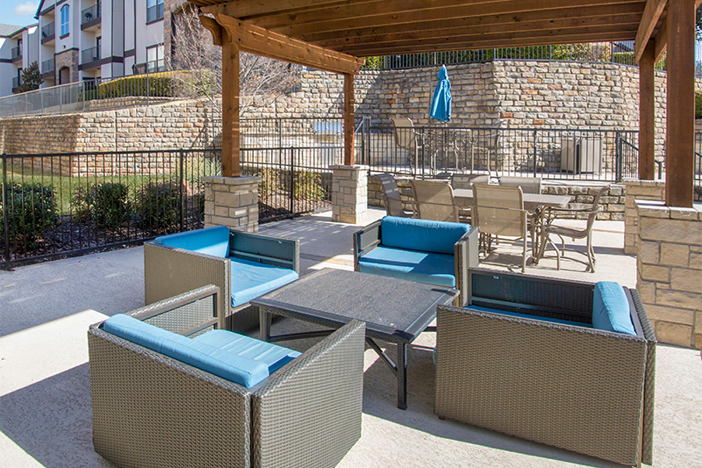 Outdoor seating area featuring modern blue cushioned chairs and a black table under a wooden pergola. A stone wall and patio furniture are visible in the background, creating a welcoming atmosphere for relaxation or socializing.
