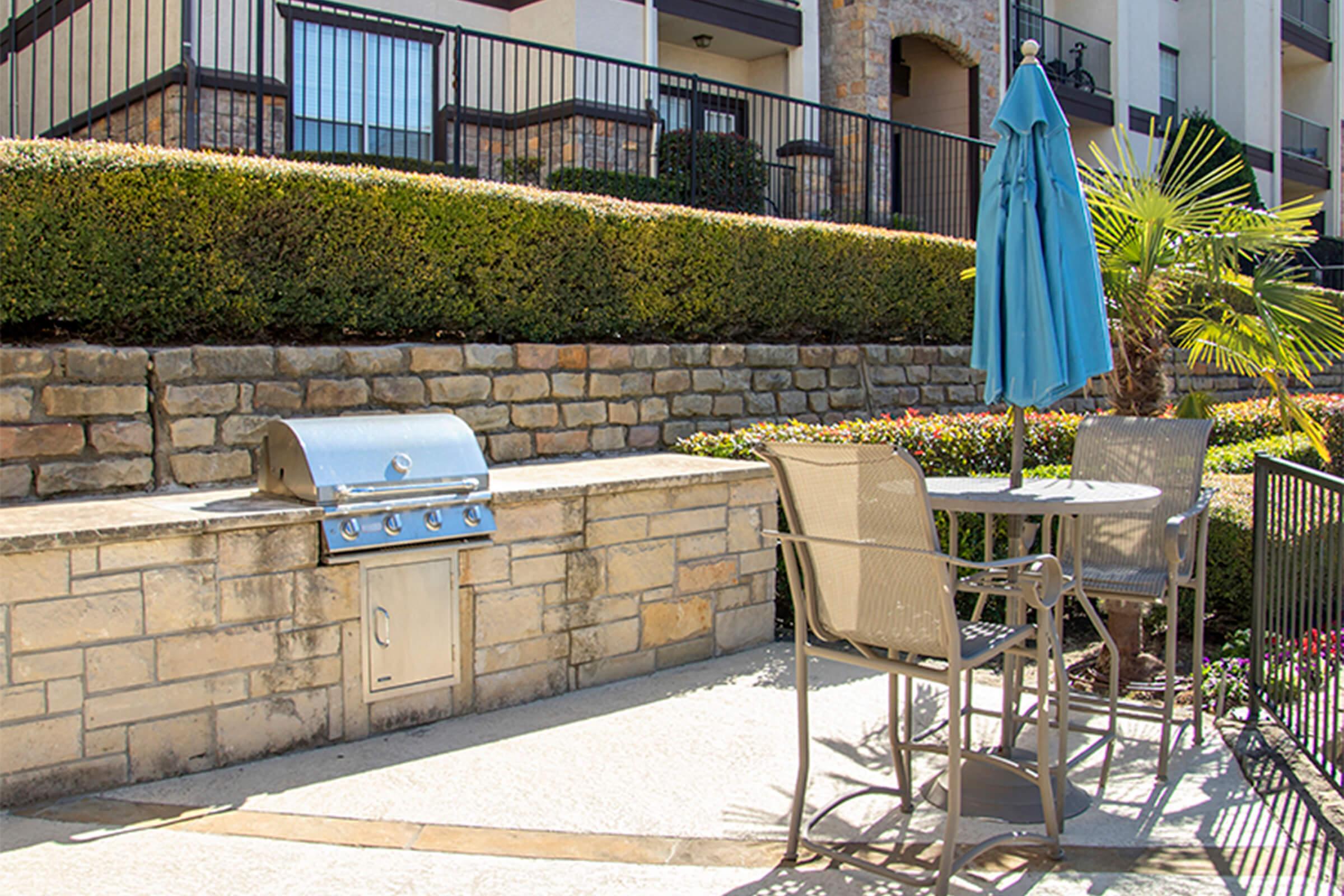 A patio area featuring a built-in barbecue grill on a stone wall, a small round table with two chairs, and a blue umbrella. Lush greenery and landscaped shrubs surround the space, creating a cozy outdoor atmosphere, ideal for dining and relaxation.