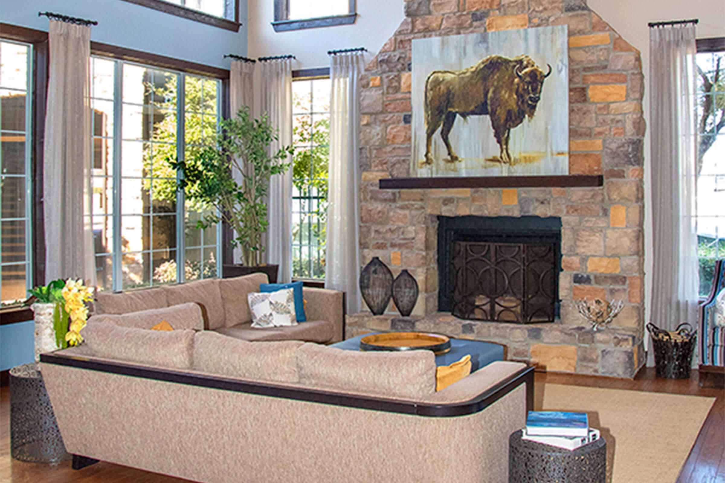 A cozy living room featuring a beige sectional sofa with colorful cushions, a large stone fireplace with a decorative screen, and a prominent bison painting above the mantel. Large windows allow natural light, complemented by potted plants and wooden decor accents.