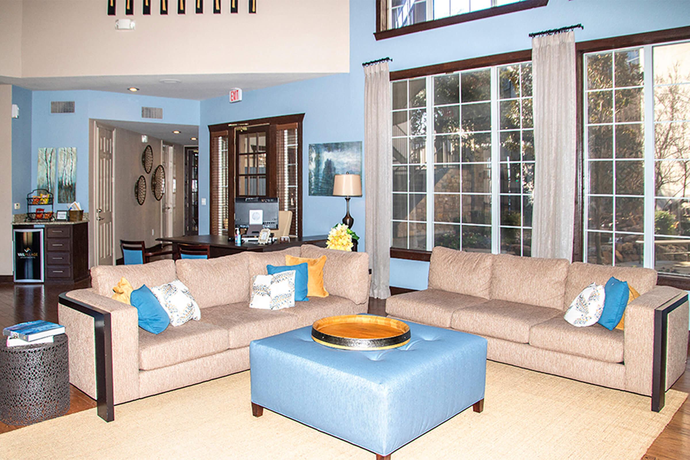 A contemporary living room featuring a light brown sectional sofa adorned with blue and white cushions. In the center, there is a blue coffee table with a round tray. Large windows allow natural light, and the walls are painted light blue. A simple desk and decorative items are visible in the background.