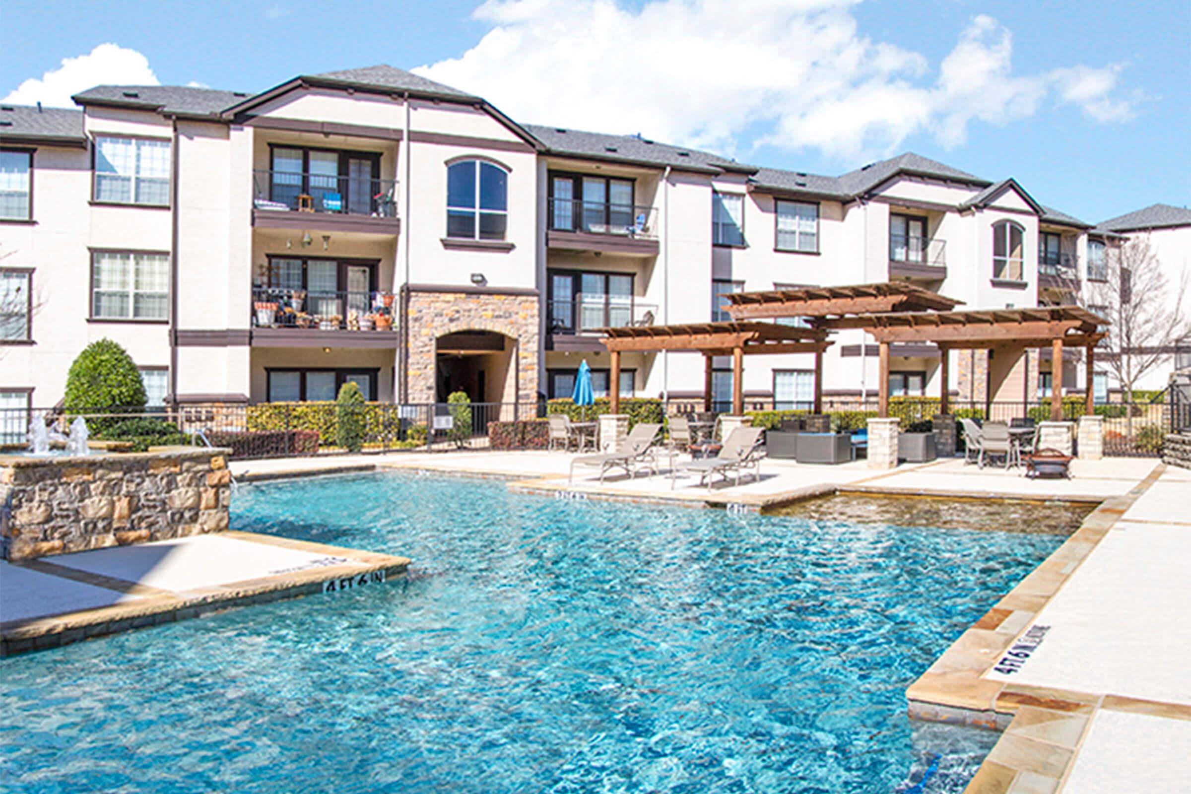A large outdoor swimming pool surrounded by lounge chairs, with several trees and shrubs nearby. In the background, there are multi-story residential buildings featuring balconies. The area is well-lit under a clear blue sky, suggesting a pleasant and inviting atmosphere.