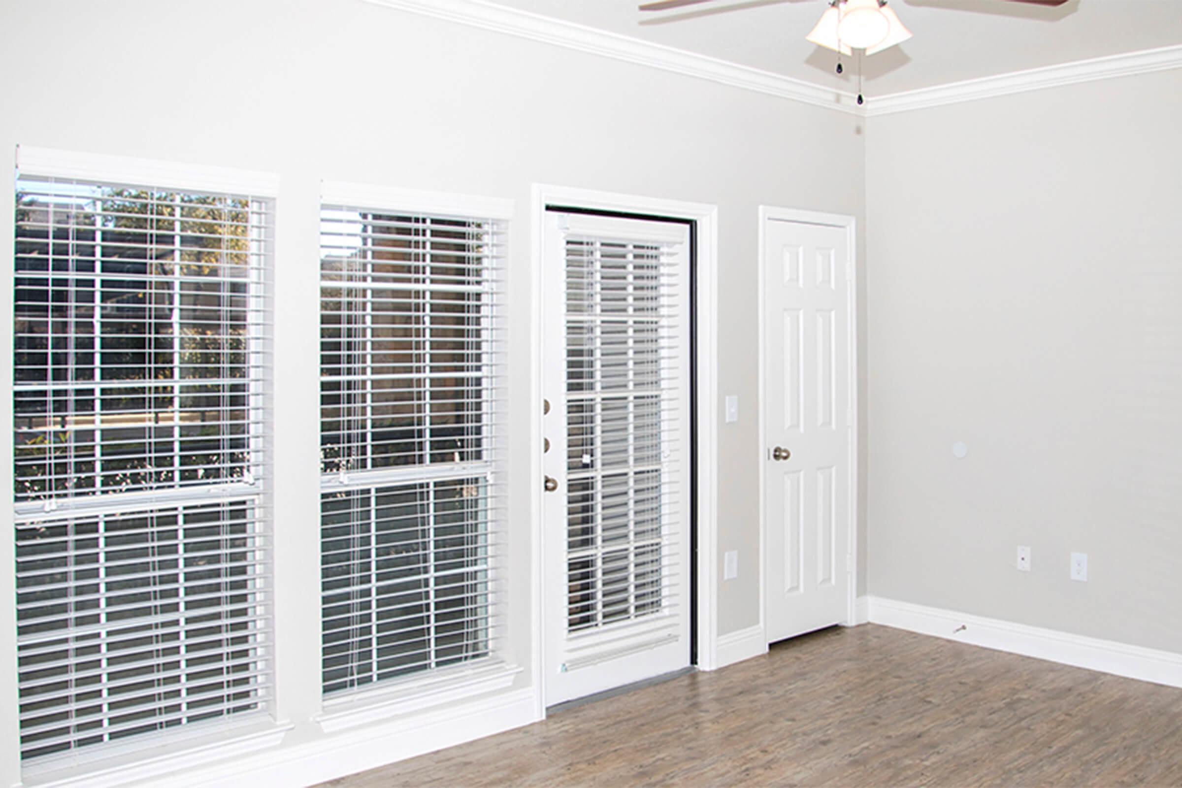 Empty room featuring light-colored walls, a ceiling fan, and large windows with white blinds. A door leads outside, and there is a closed door on the right. The floor has a wood-like finish, creating a bright and airy atmosphere.