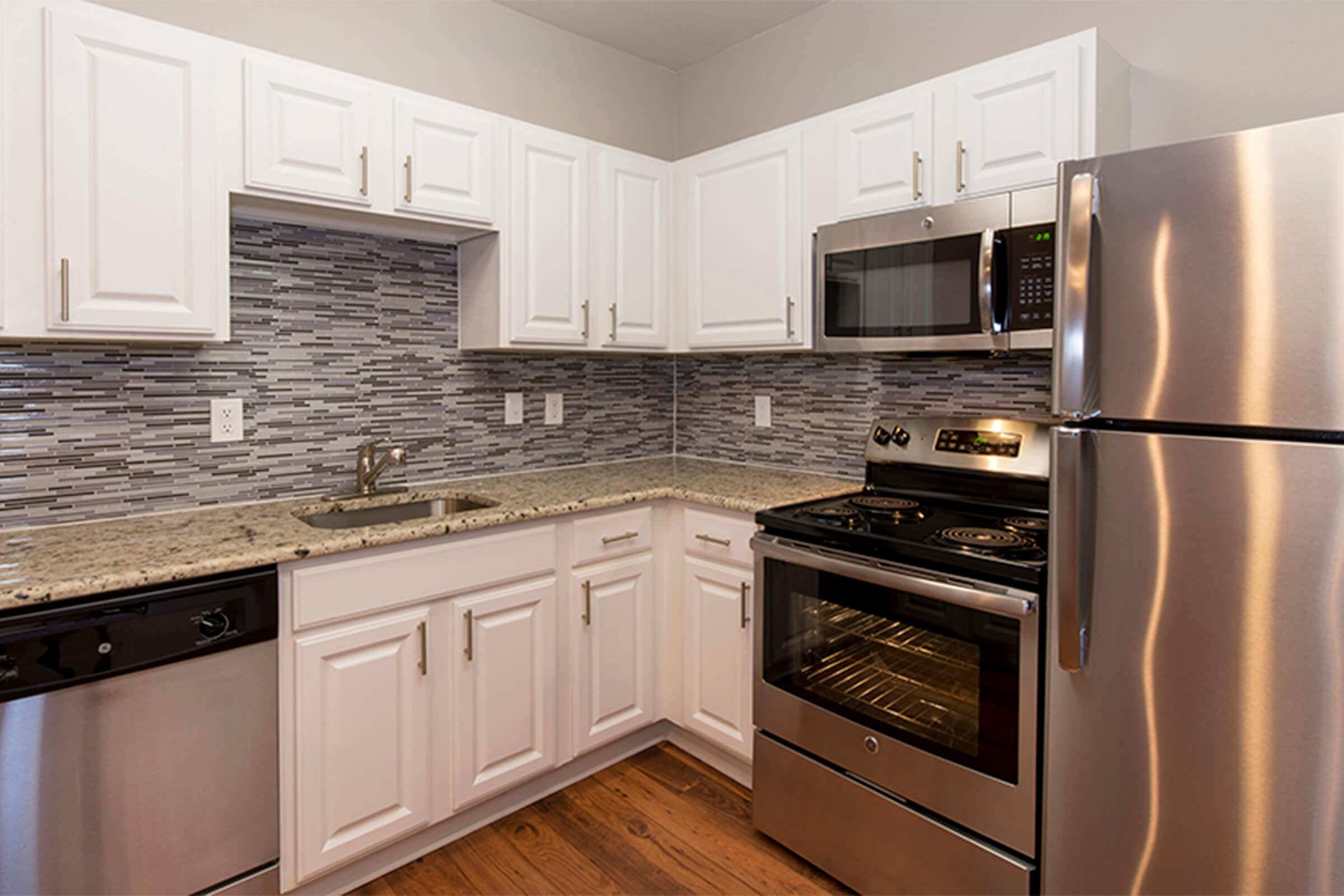 Modern kitchen with white cabinets, granite countertop, stainless steel appliances including an oven, microwave, and dishwasher. A tiled backsplash complements the design, and there is a sink in the corner. The overall aesthetic is clean and contemporary, with warm wooden flooring.