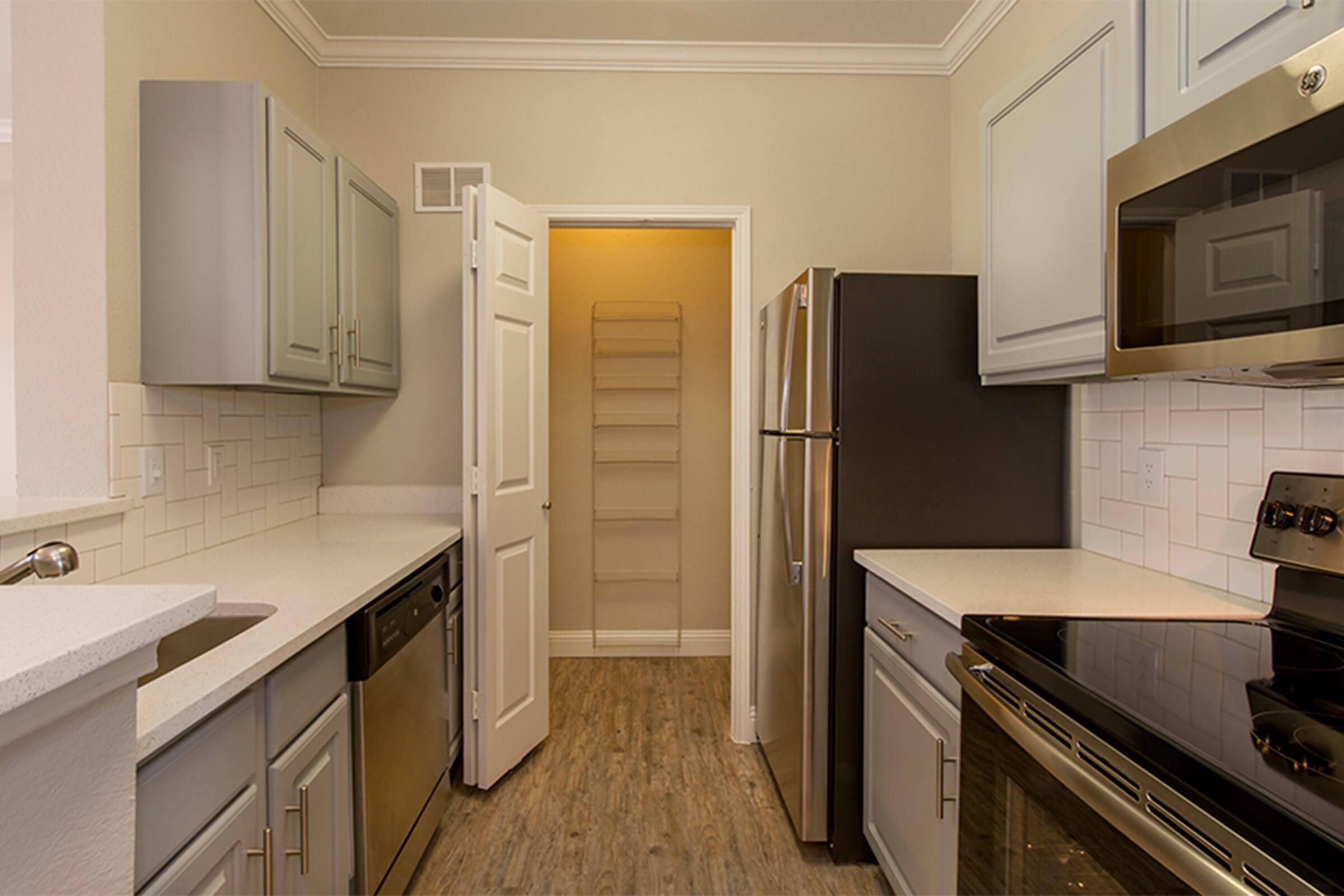 A modern kitchen featuring light gray cabinets, a stainless steel dishwasher, and an electric stove. The countertops are white, and there's a spacious feel with wood-like flooring. A door leads to a pantry or utility area, adding functionality to the design.