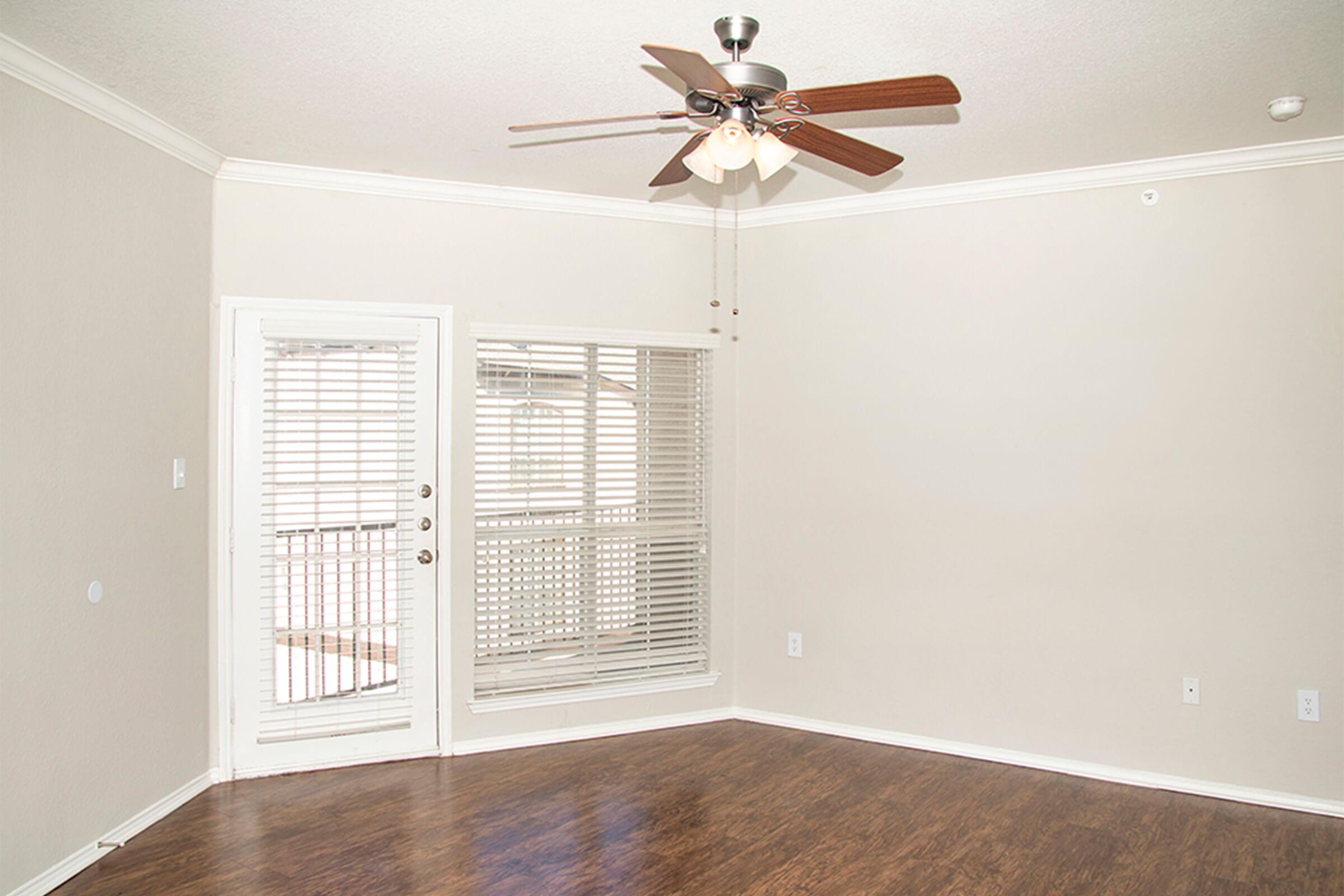Empty room with light-colored walls and a ceiling fan. A sliding glass door with vertical blinds leads to a balcony. The floor is made of dark wood. Natural light comes in from outside, creating a spacious and bright atmosphere.