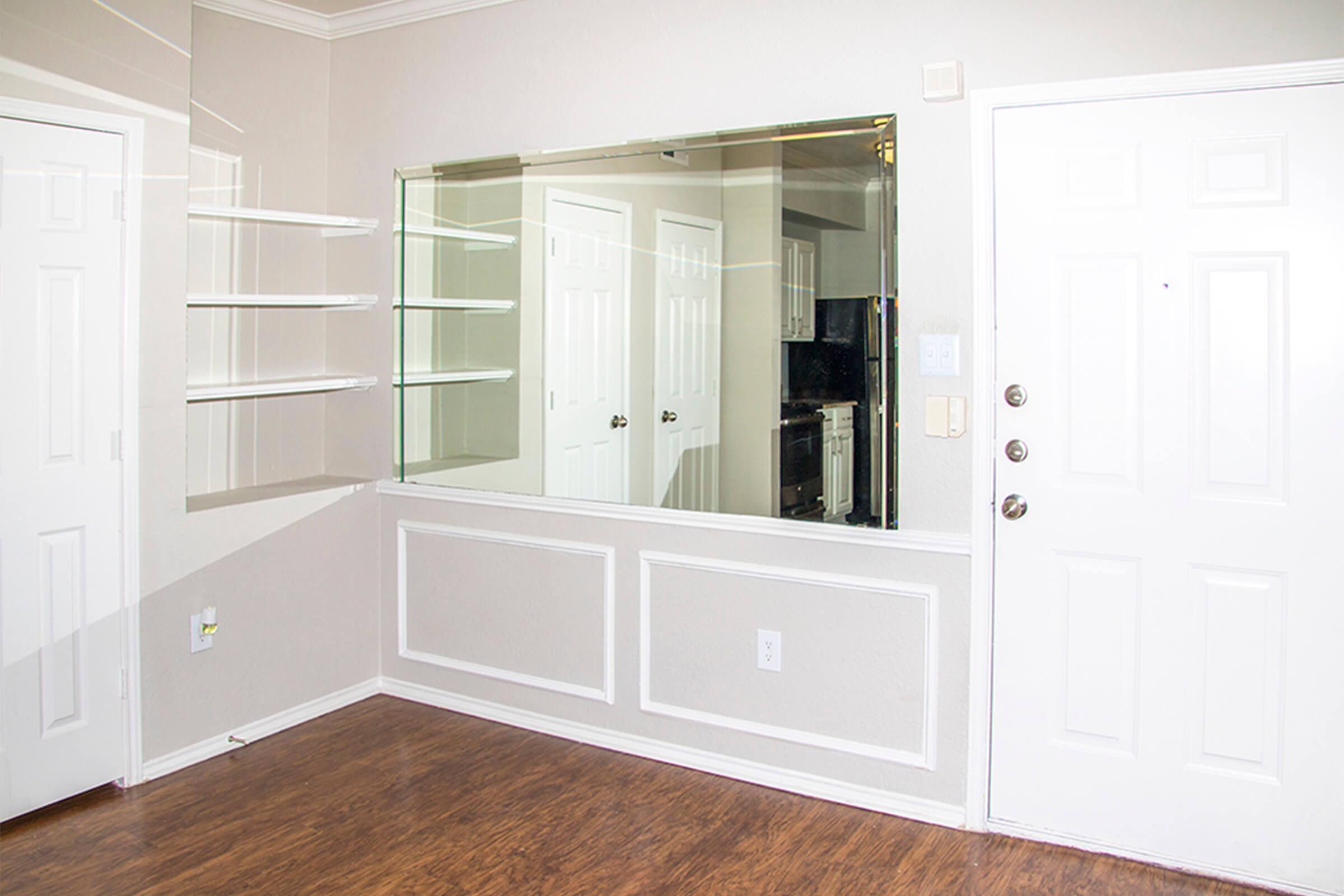 A well-lit room featuring a light-colored wall with a large mirror and open shelving. The entrance door is visible on the right, while the left side shows another door. The floor is made of wood. The room has a clean and simple aesthetic, suitable for various design styles.