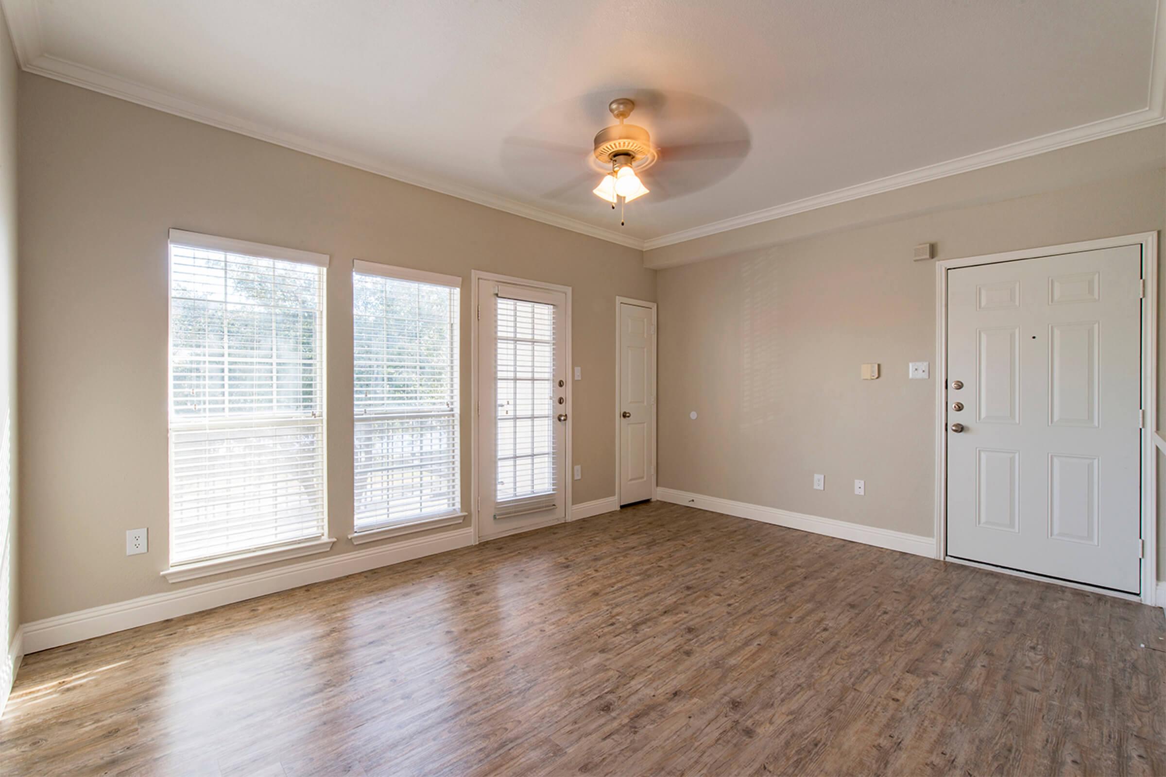 A bright, unfurnished living room featuring light beige walls, large windows with blinds, and a ceiling fan. There are two doors, one leading outside and the other to a small closet. The floor is a warm wood laminate, and the overall ambiance is open and inviting.