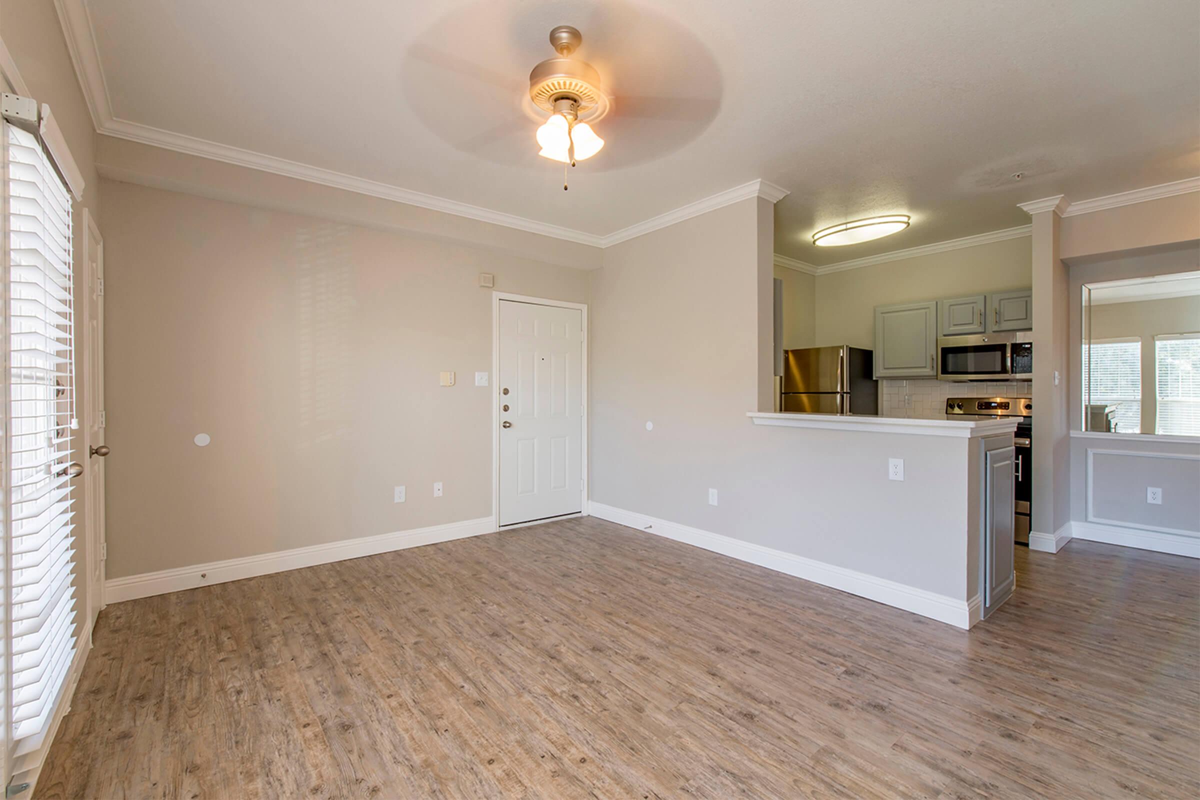 Spacious living area with light-colored walls and wood-like flooring. Features a ceiling fan and an open layout leading to a kitchen with modern appliances. A front door is visible, and large windows allow natural light to brighten the space.