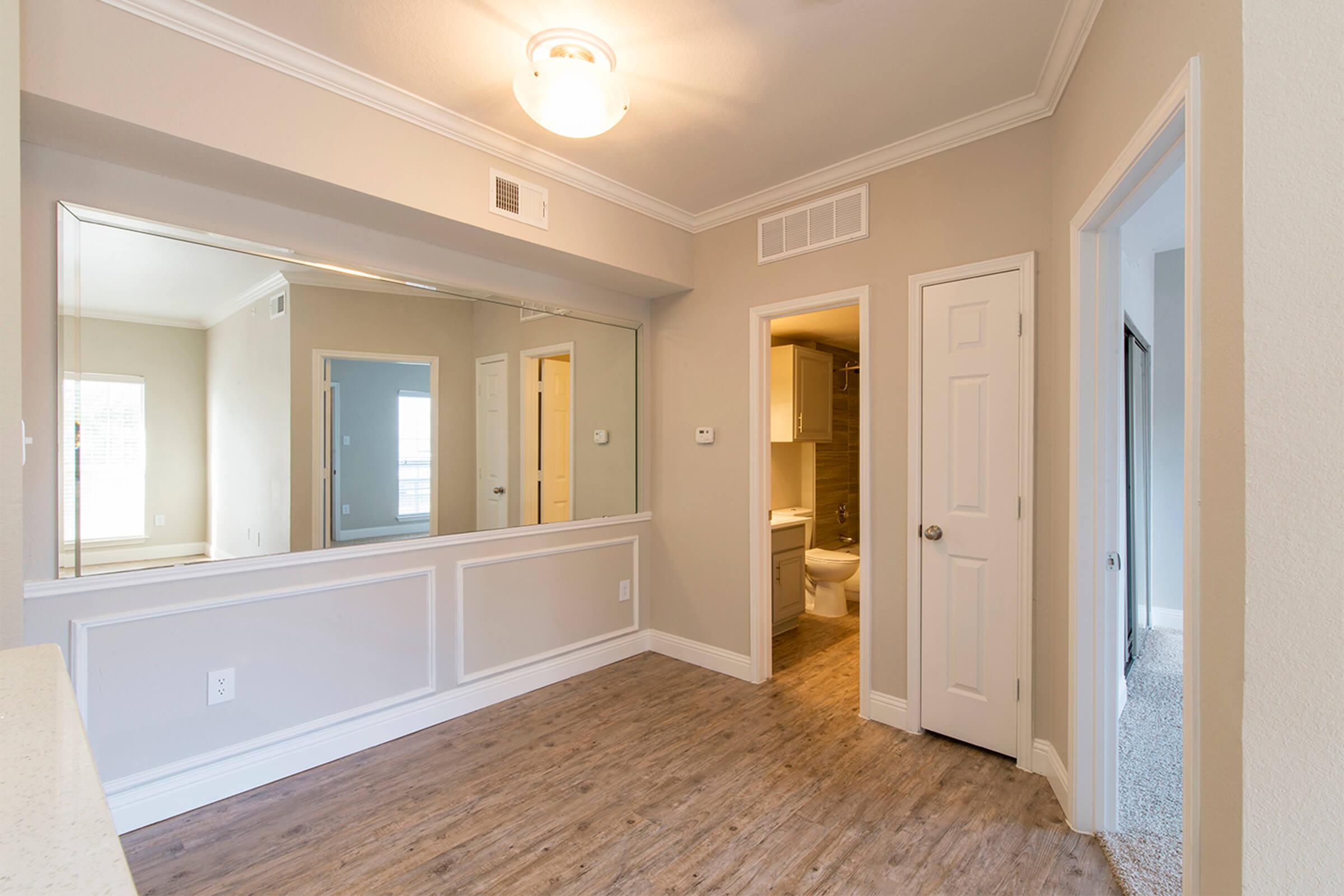 Empty interior of a room featuring a large mirror, light-colored walls, and wood-like flooring. A doorway on the right leads to a bathroom, while another doorway on the left opens to an adjacent space. Natural light brightens the room, enhancing its spacious appearance.