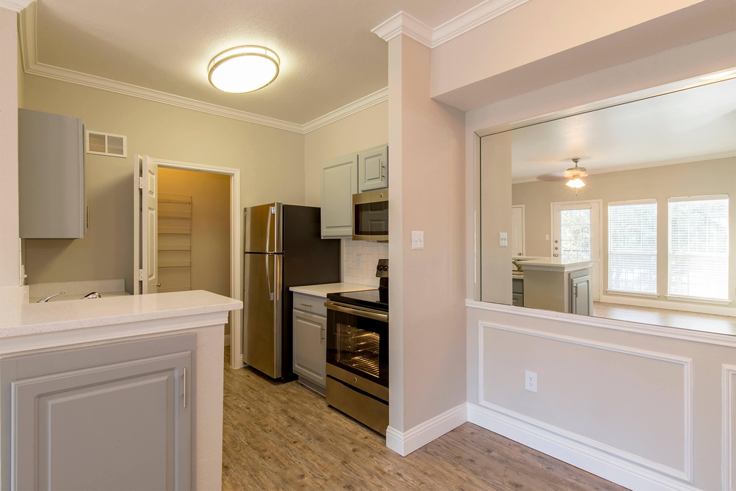 Modern kitchen featuring light-colored cabinetry, stainless steel appliances, and a spacious layout. Includes a refrigerator, oven, and a view of a living area through a cut-out window. The walls are painted in neutral tones, and there is ample natural light from nearby windows.