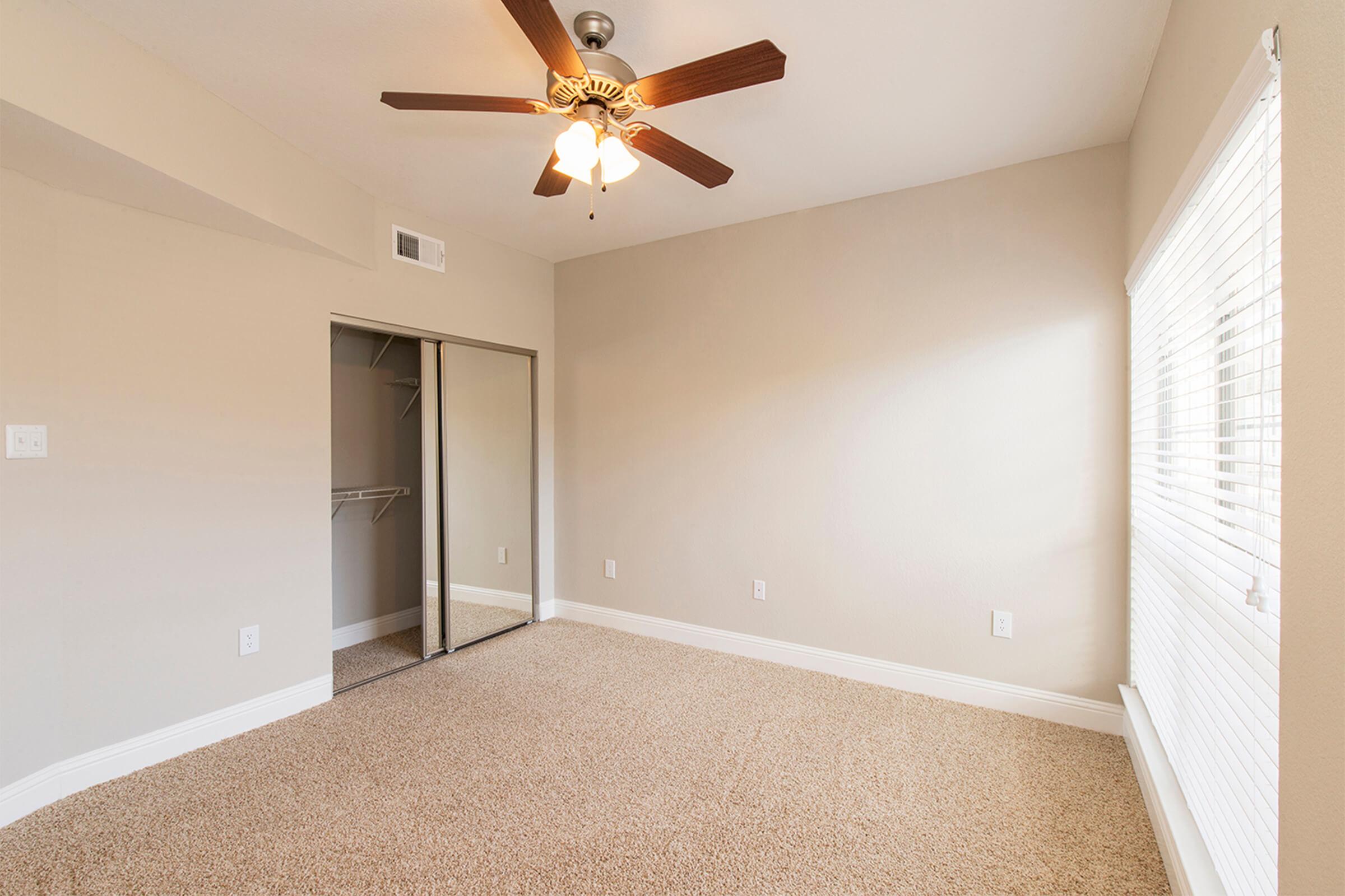 A clean, well-lit bedroom featuring a ceiling fan, beige walls, and carpeted flooring. There is a closet with sliding mirrored doors on one side, and a window with blinds allowing natural light to enter. The space appears inviting and ready for personalization.