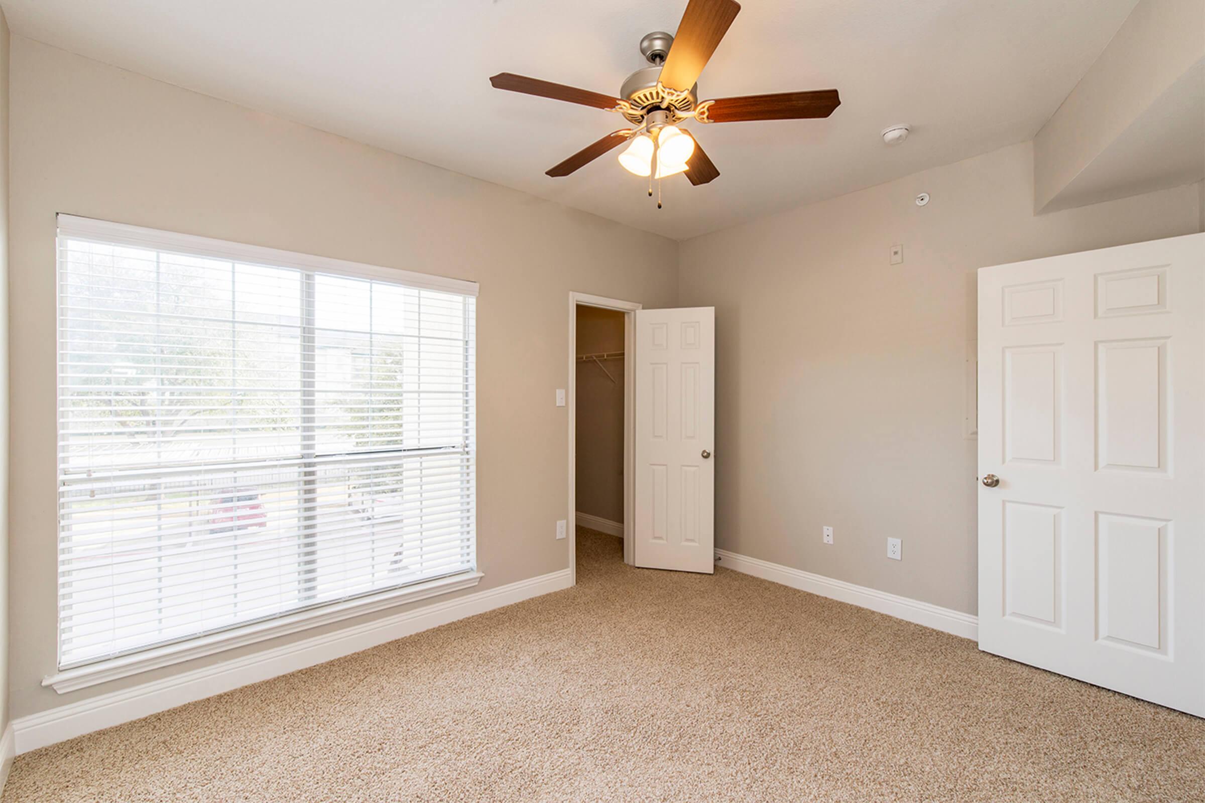 Bright and airy bedroom featuring beige walls and plush carpeting. A ceiling fan with wooden blades hangs from the ceiling, and large windows with white blinds provide natural light. There is a closet door to the left and a white door on the right, adding to the room's open and inviting layout.