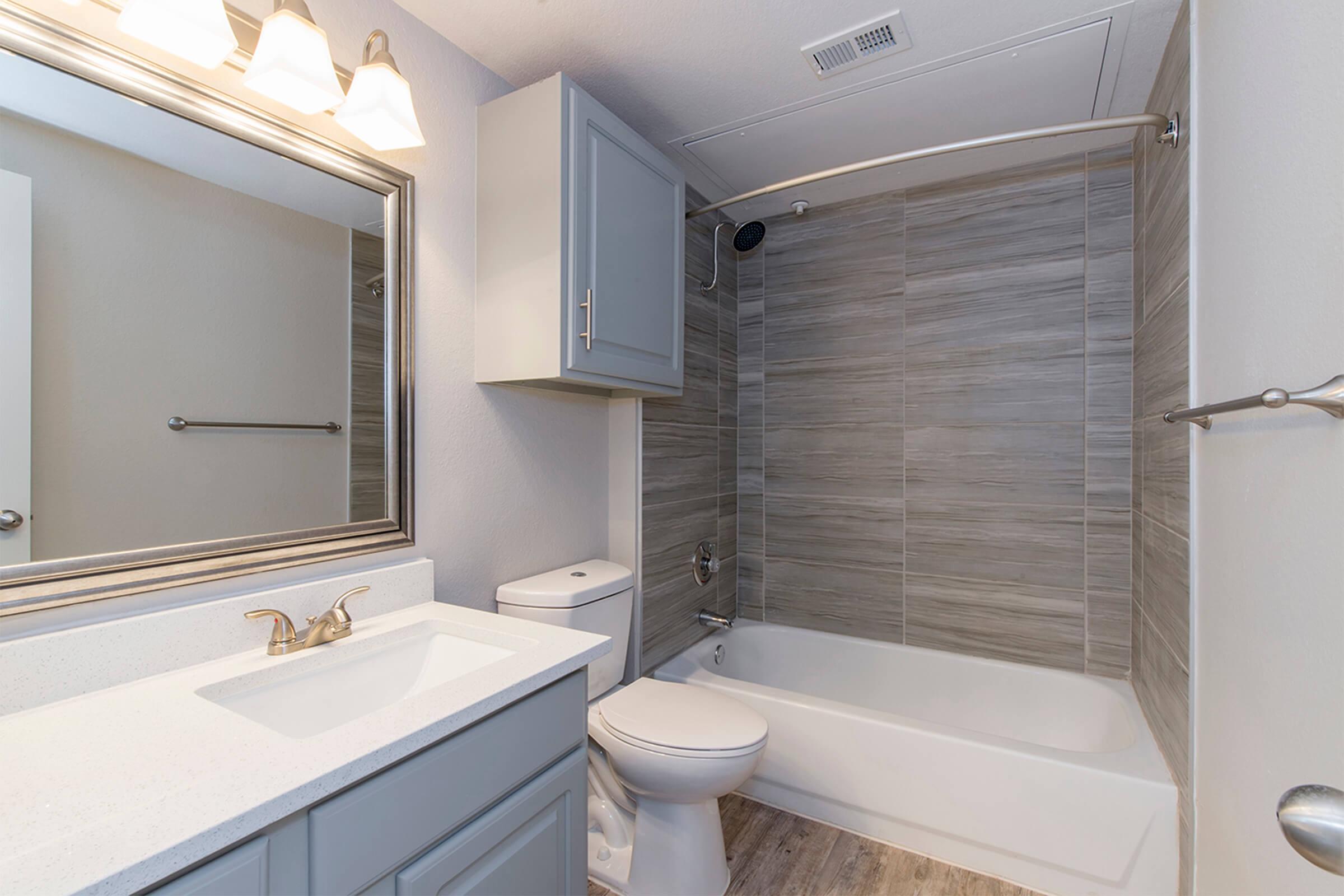 A modern bathroom featuring a light gray wall with a bathtub and shower combination. There is a white toilet next to the bathtub, and a stylish vanity with a sink and mirror above it. Soft lighting from overhead fixtures enhances the clean, contemporary look of the space.
