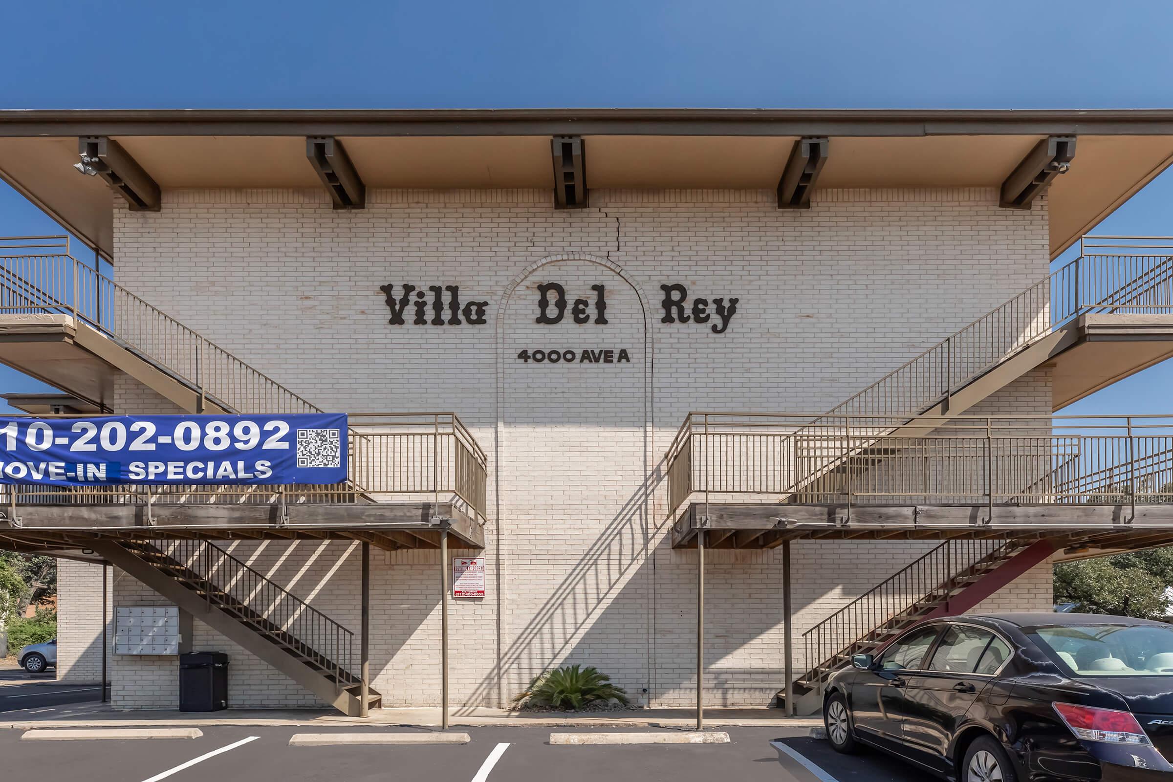 Exterior view of a three-story apartment building with a sign reading "Villa Del Rey" and the address "4000 Aveh." Stairs on both ends lead to upper levels. A parking lot is visible in front, featuring several parked cars and a banner advertising move-in specials. Clear blue sky overhead.
