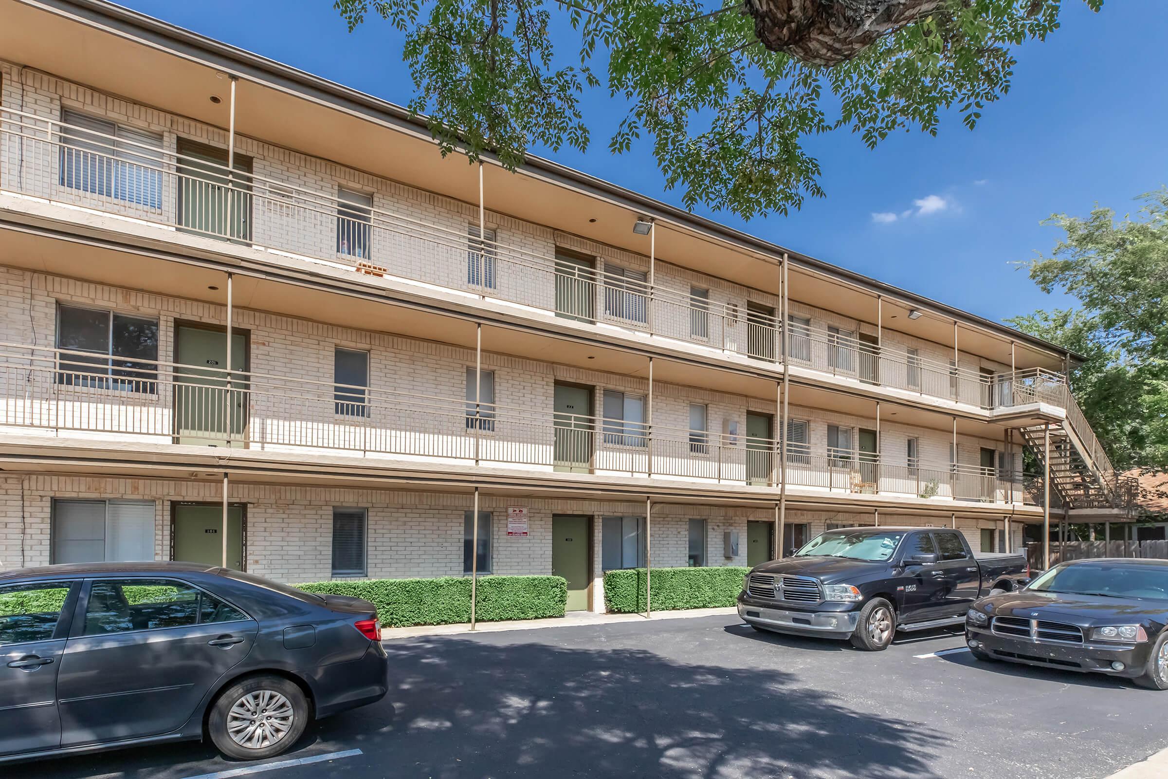 Two-story apartment building with a brick exterior, featuring multiple balconies and green doors. Several parked cars are visible in the foreground, and there are trees nearby under a clear blue sky.