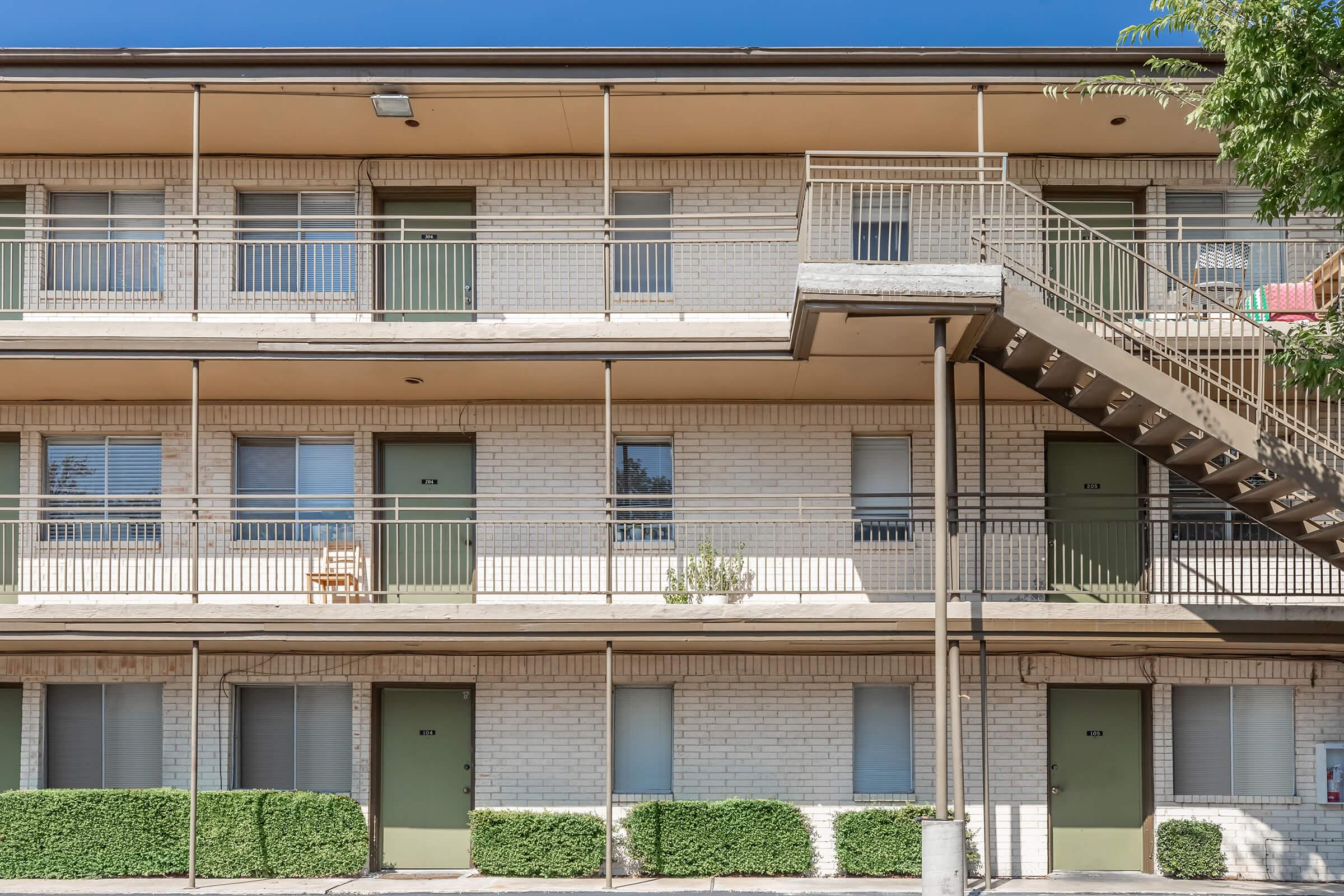 A multi-story building featuring a light-colored exterior, multiple green doors, and a staircase on the right side. There are several windows with curtains, and a single wooden chair is visible on a balcony. The landscape includes neatly trimmed shrubs at the base of the building.
