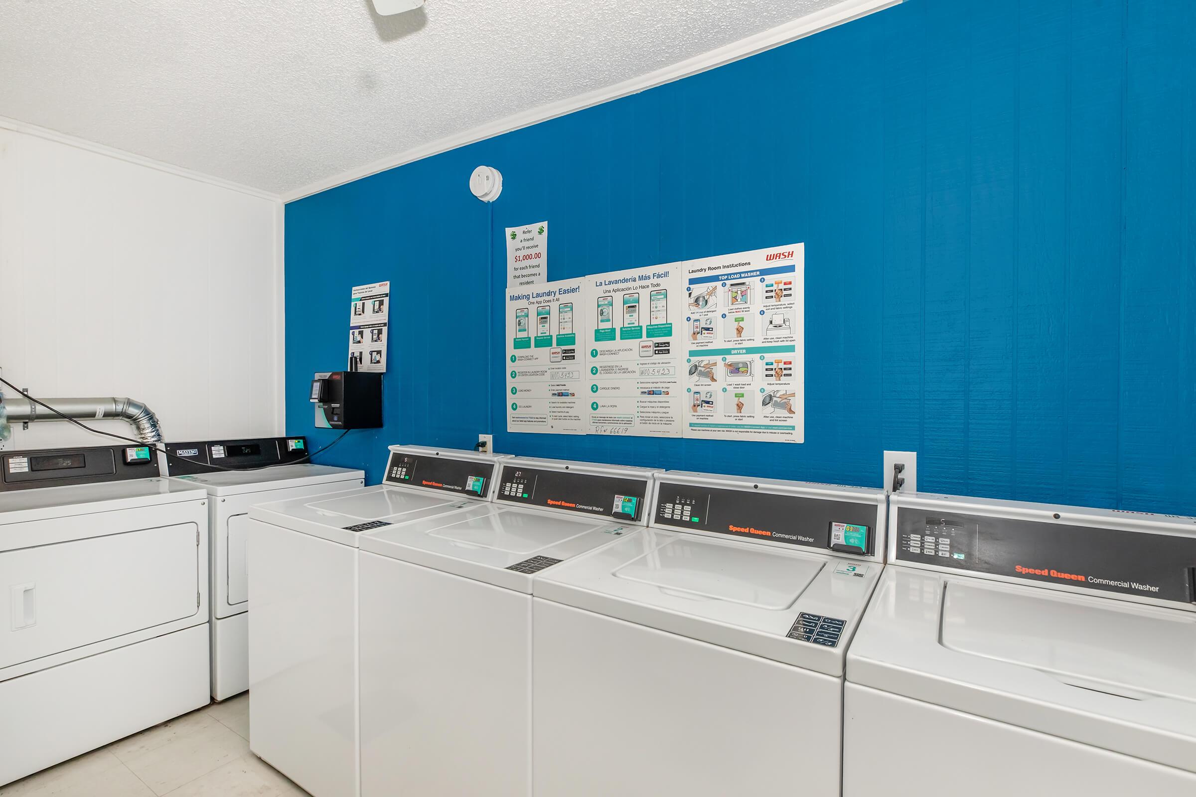 A laundry room featuring several white washing machines and dryers against a blue wall. A chart with laundry instructions and equipment usage is displayed above the machines. The room is clean and functional, designed for communal laundry use.