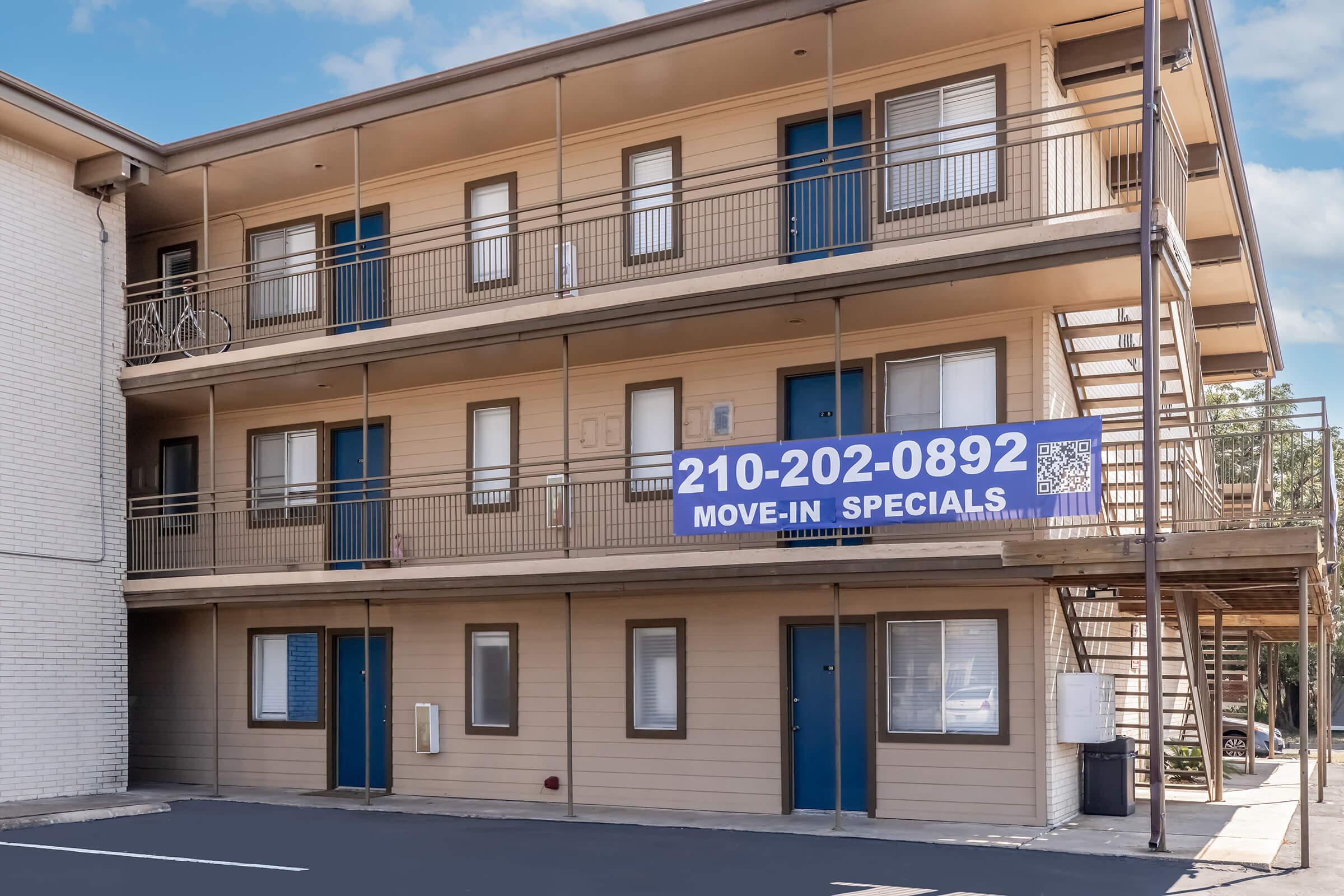 Exterior view of a two-story apartment building featuring multiple balconies and a staircase. A large "MOVE-IN SPECIALS" banner is displayed prominently on the front, along with a contact number. The building has a light-colored exterior with blue doors and windows. The surrounding area includes a paved parking lot.