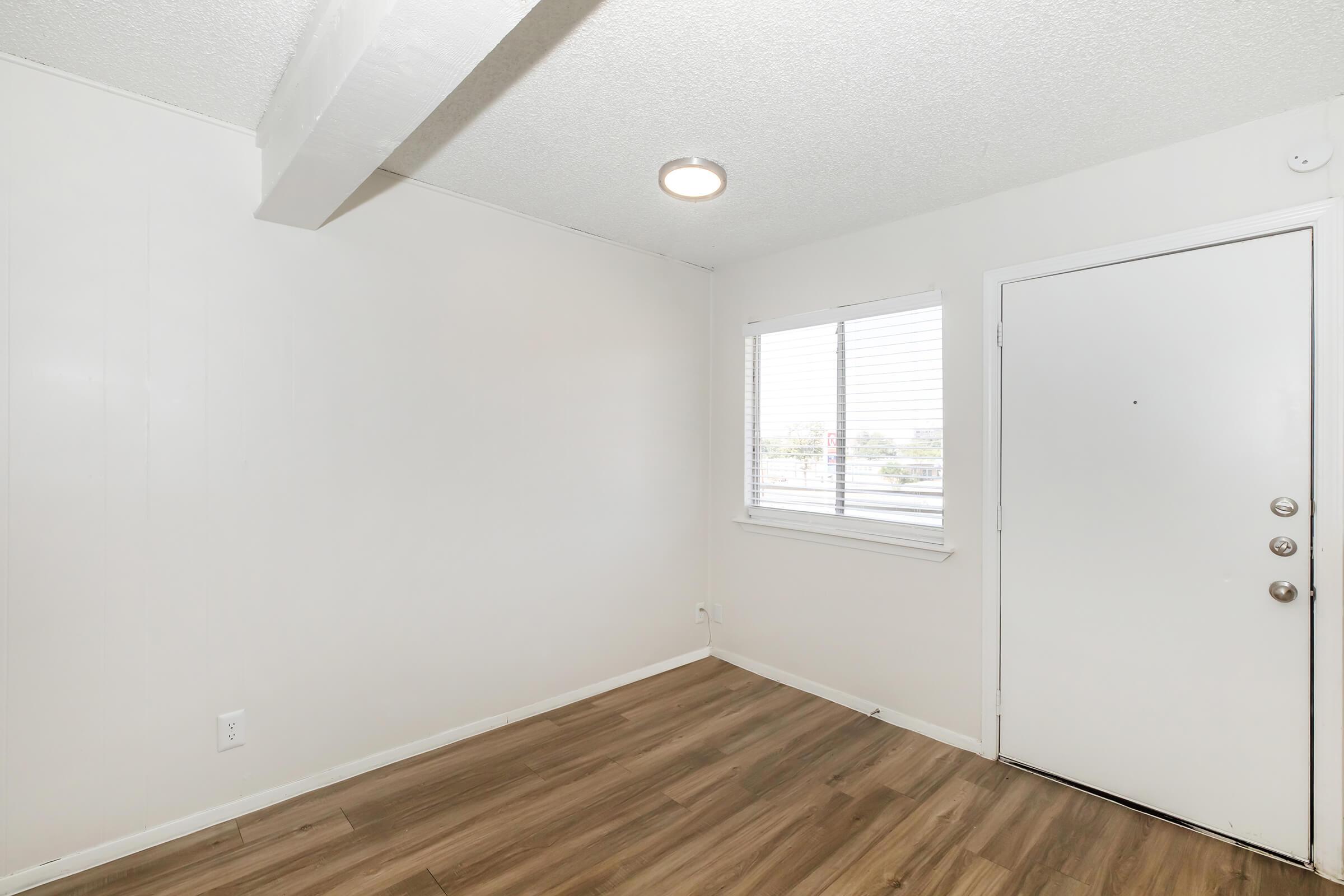 A well-lit, empty room with light-colored walls, a window featuring blinds, and a door on one side. The floor is covered in laminate wood. A ceiling light fixture is visible, and there are no furnishings in the space, giving it a clean and minimalist appearance.