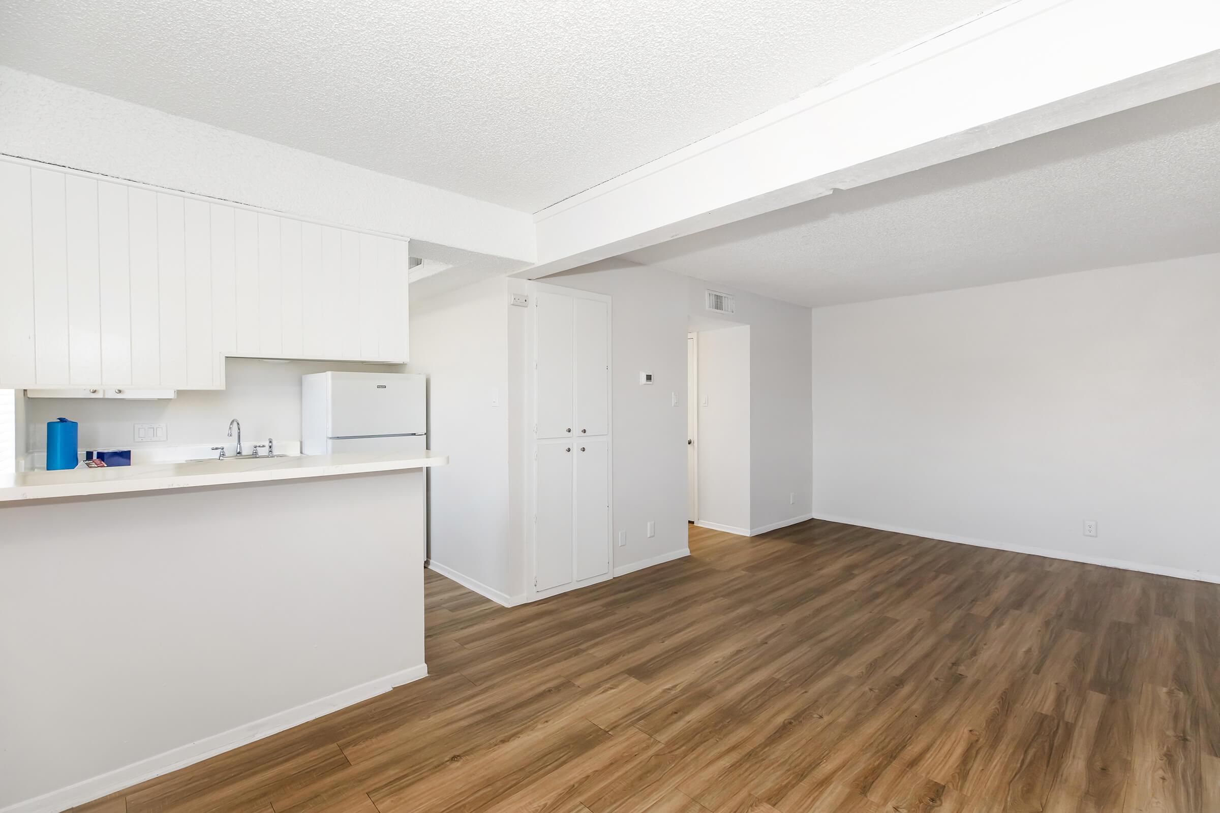 Interior of a modern apartment featuring an open floor plan. The kitchen is visible on the left with white cabinets and a refrigerator. The living area has light-colored walls and a wooden floor. Natural light enters from a window, creating a spacious and inviting atmosphere.