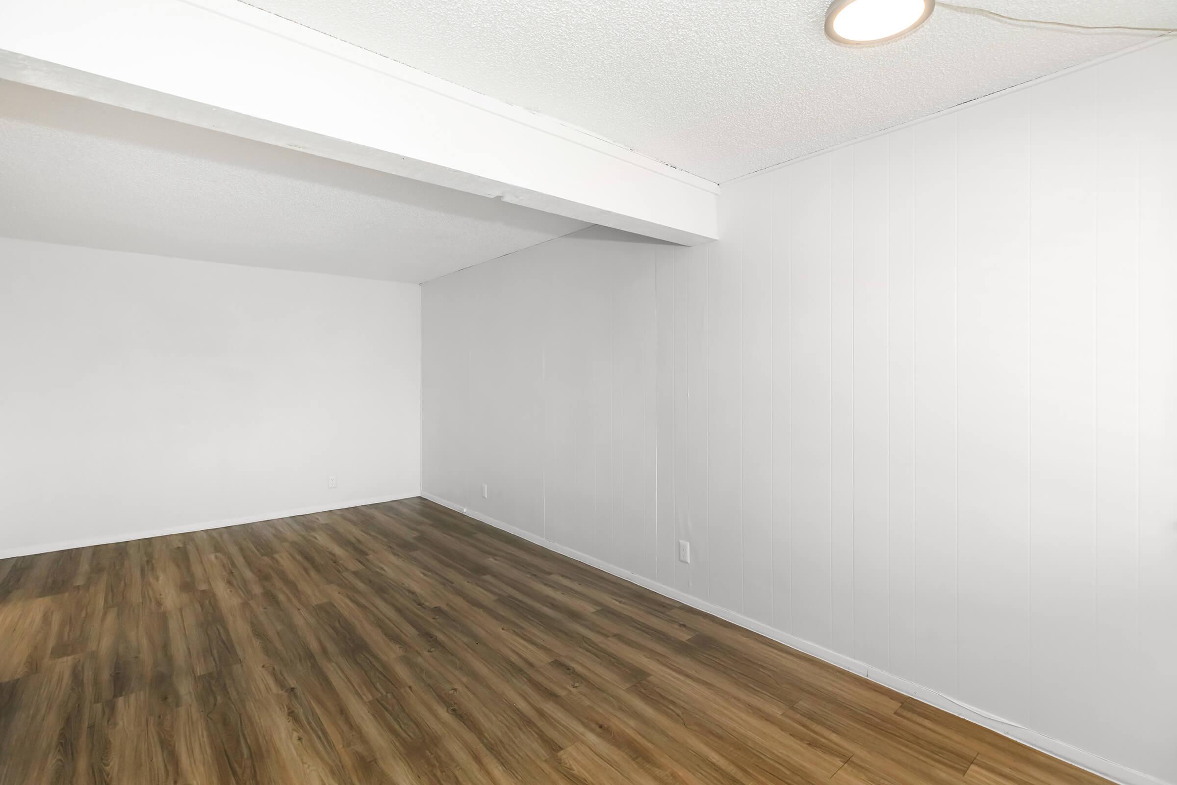 A bright, empty room with light-colored walls and a smooth wooden floor. There is a single ceiling light fixture visible. The room appears spacious with minimal furnishings, giving it an airy feel.
