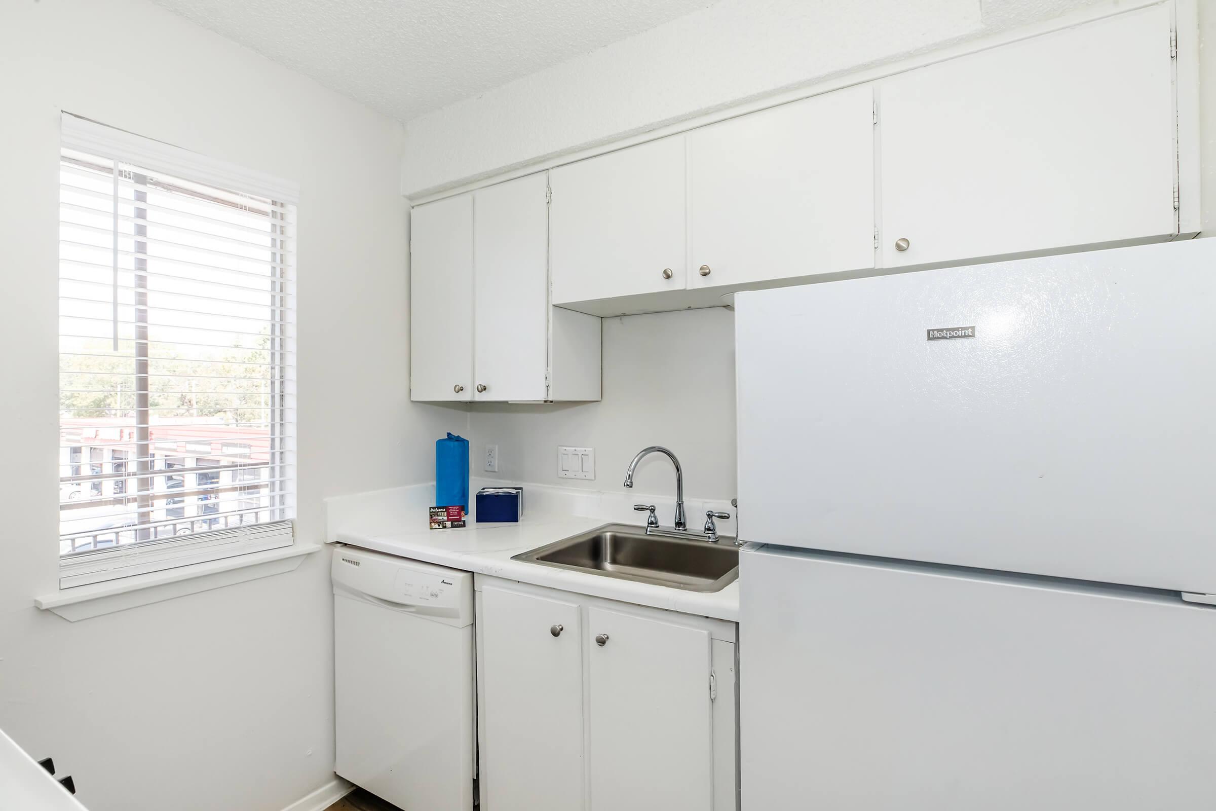 A small modern kitchen featuring white cabinetry, a stainless steel sink, a dishwasher, and a refrigerator. A window with blinds allows natural light to enter, and there are minimal decorative elements, creating a clean and functional space.