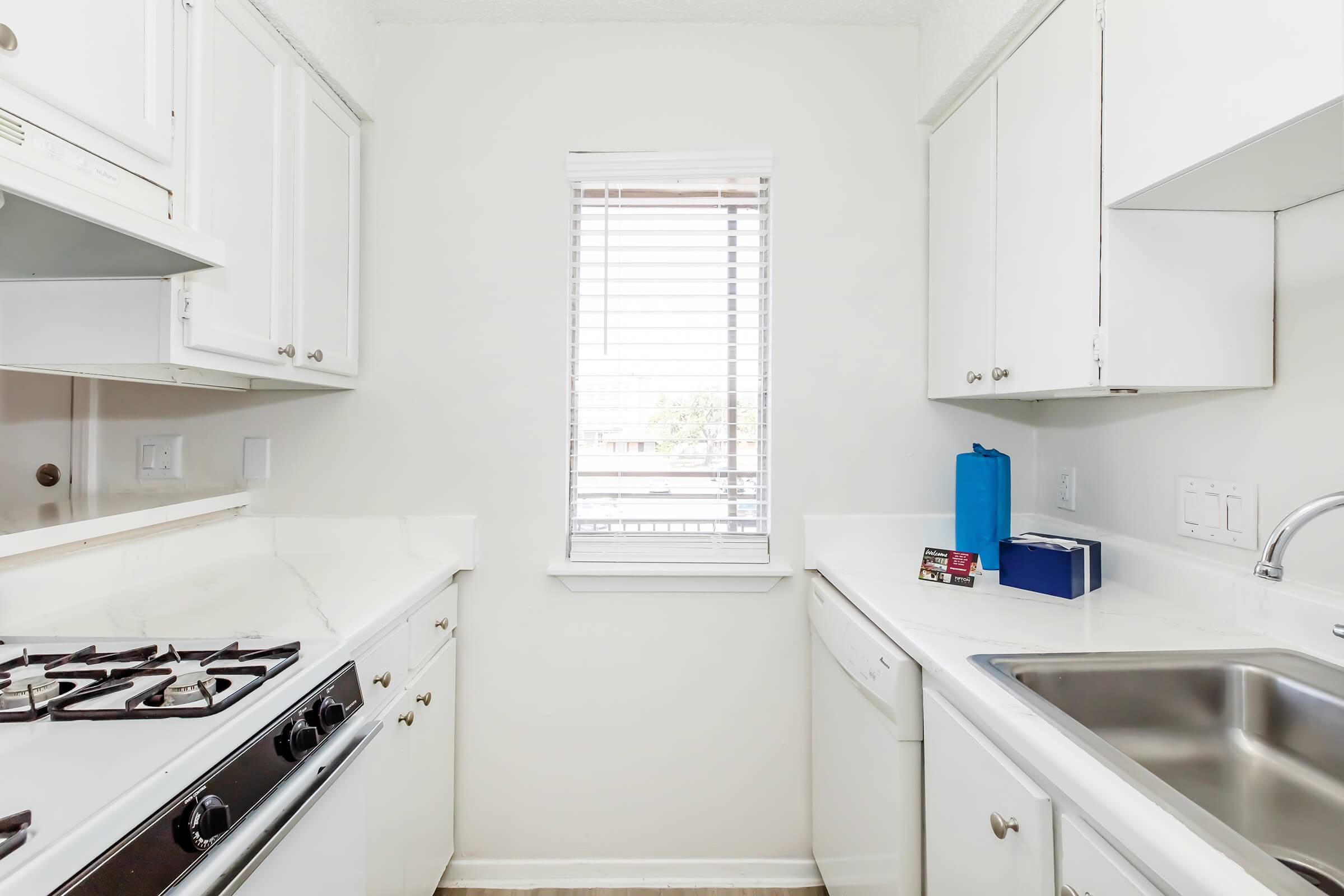 A small, modern kitchen featuring white cabinetry, a gas stove, a sink, and a window with blinds. The countertops are also white, and there are a few kitchen appliances like a blue container and a box on the counter. Natural light enters through the window, creating a bright atmosphere.
