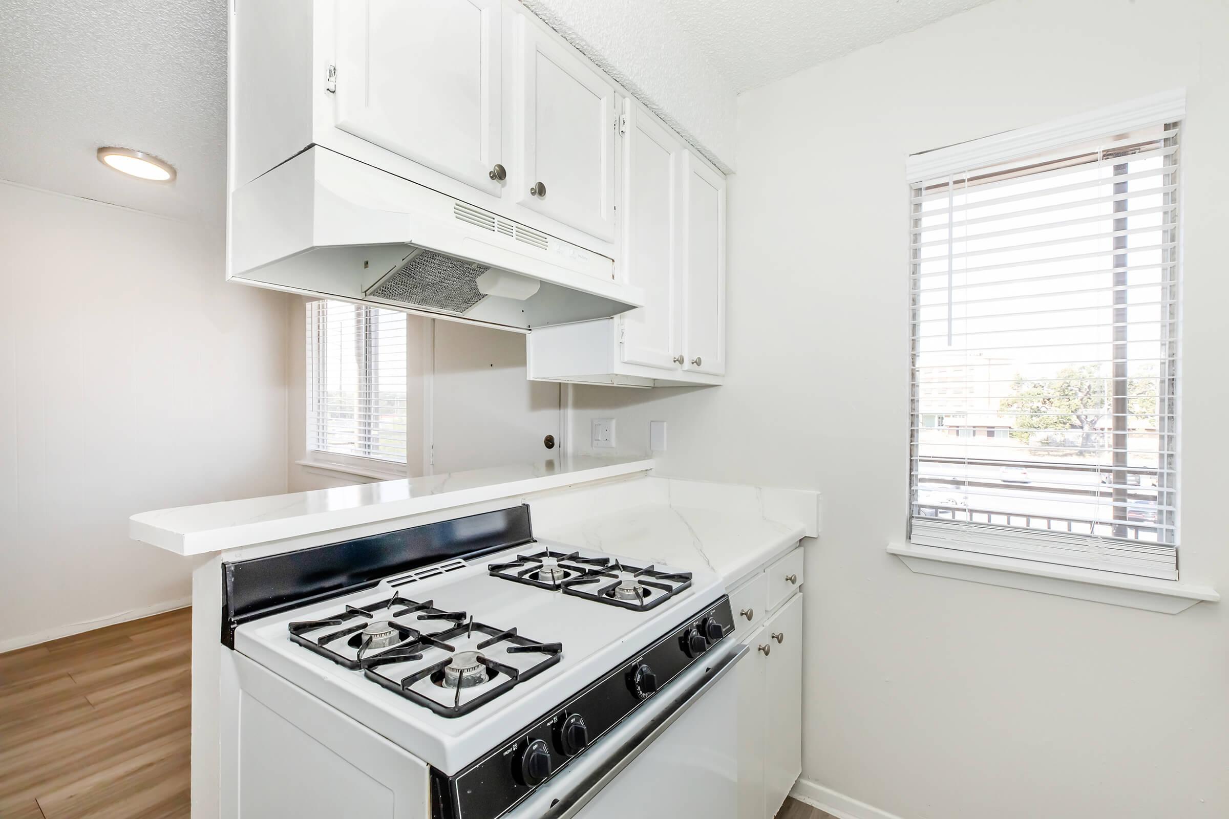 A modern kitchen featuring a white gas stove with multiple burners, a sleek countertop, and a range hood. Natural light streams in through a nearby window with horizontal blinds, illuminating the clean design and neutral color palette of the space.