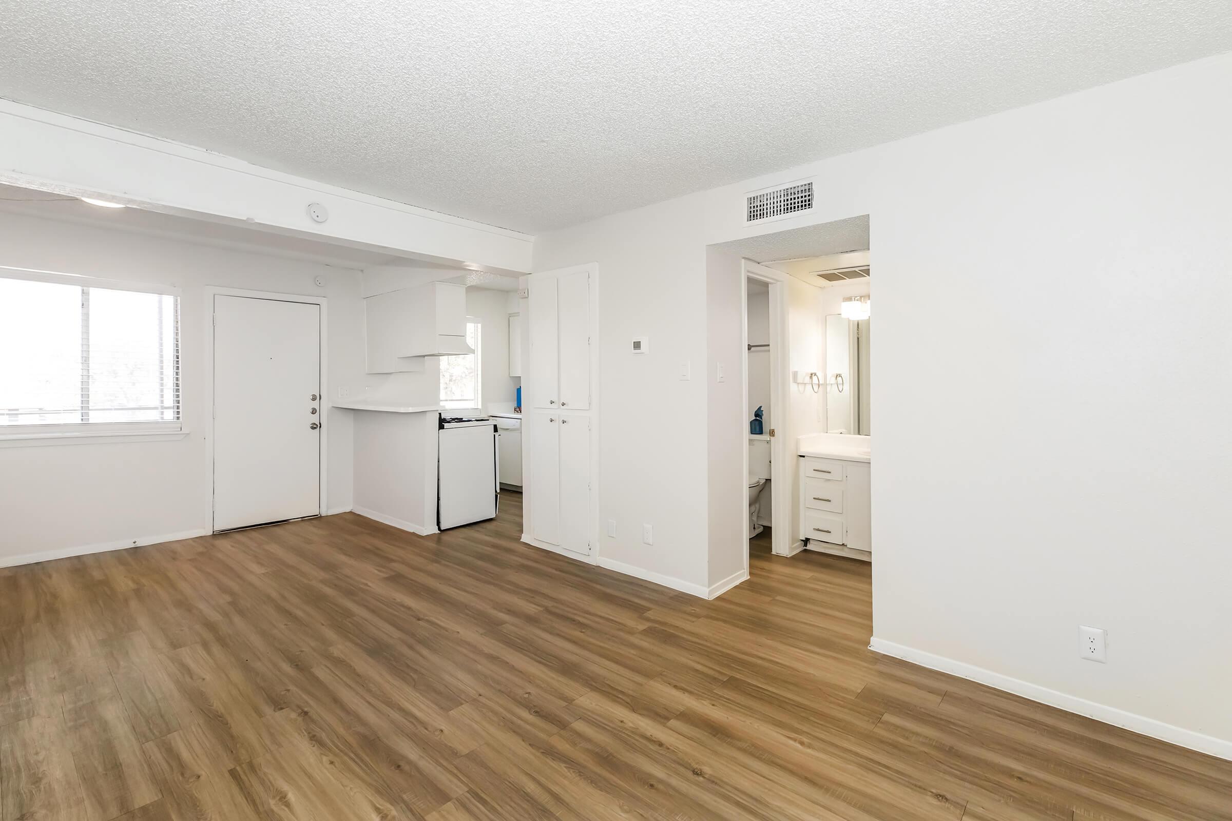 A spacious, well-lit living area featuring light wood flooring, a kitchenette on the left with a white countertop and appliances, and a doorway leading to a bathroom on the right. Large windows provide natural light, and the walls are painted a neutral color, creating a clean and modern atmosphere.