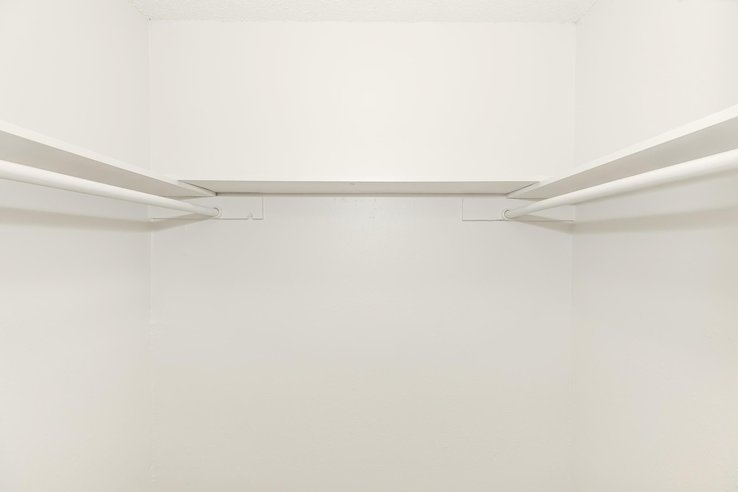 Empty white closet interior with two horizontal clothing rails attached to the walls, featuring a plain, unadorned backdrop. The lighting is bright, creating a clean and minimalistic appearance.