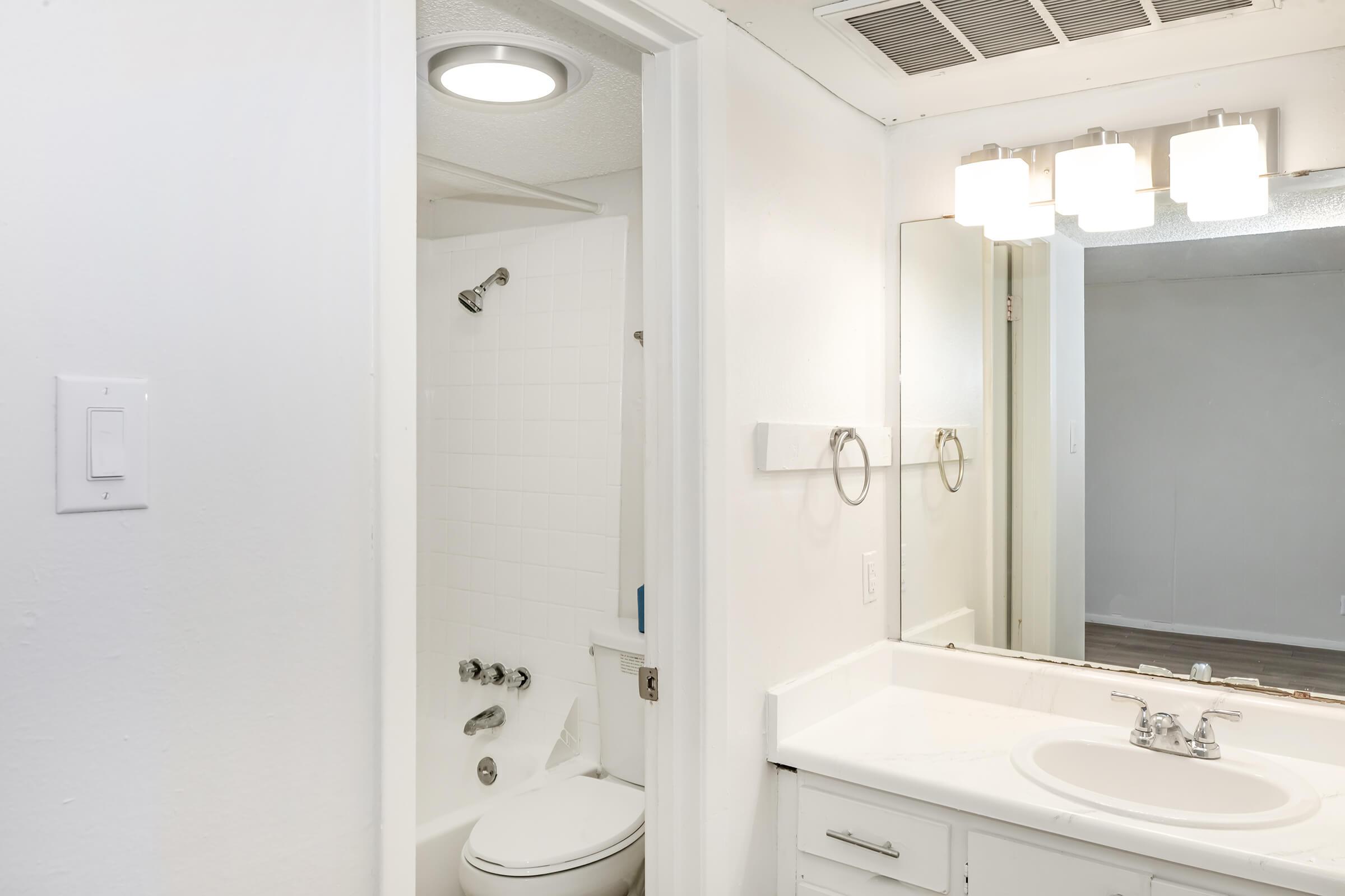 A clean, modern bathroom featuring a white vanity with a sink, a large mirror with three light fixtures above, a small shower area with a glass door, and a toilet. The walls are painted white, and the flooring is a light wood finish, creating a bright and minimalist aesthetic.