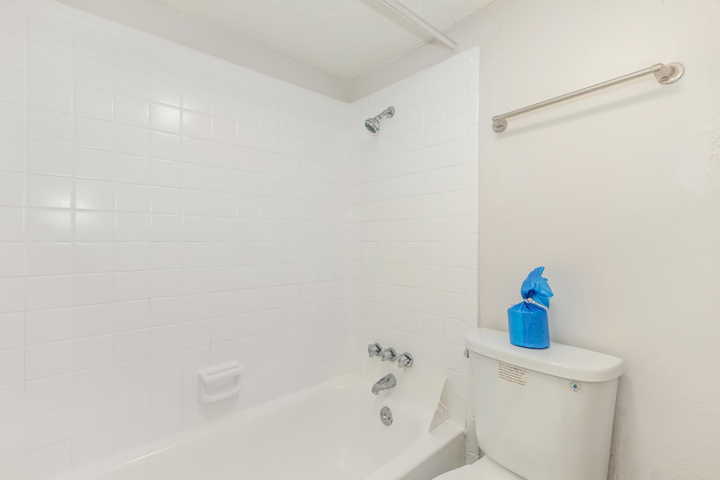 A clean bathroom featuring a white tiled bathtub with a showerhead, a toilet beside it, and a blue bag placed on the toilet tank. The walls and fixtures are bright and minimalistic, providing a fresh and simple aesthetic.